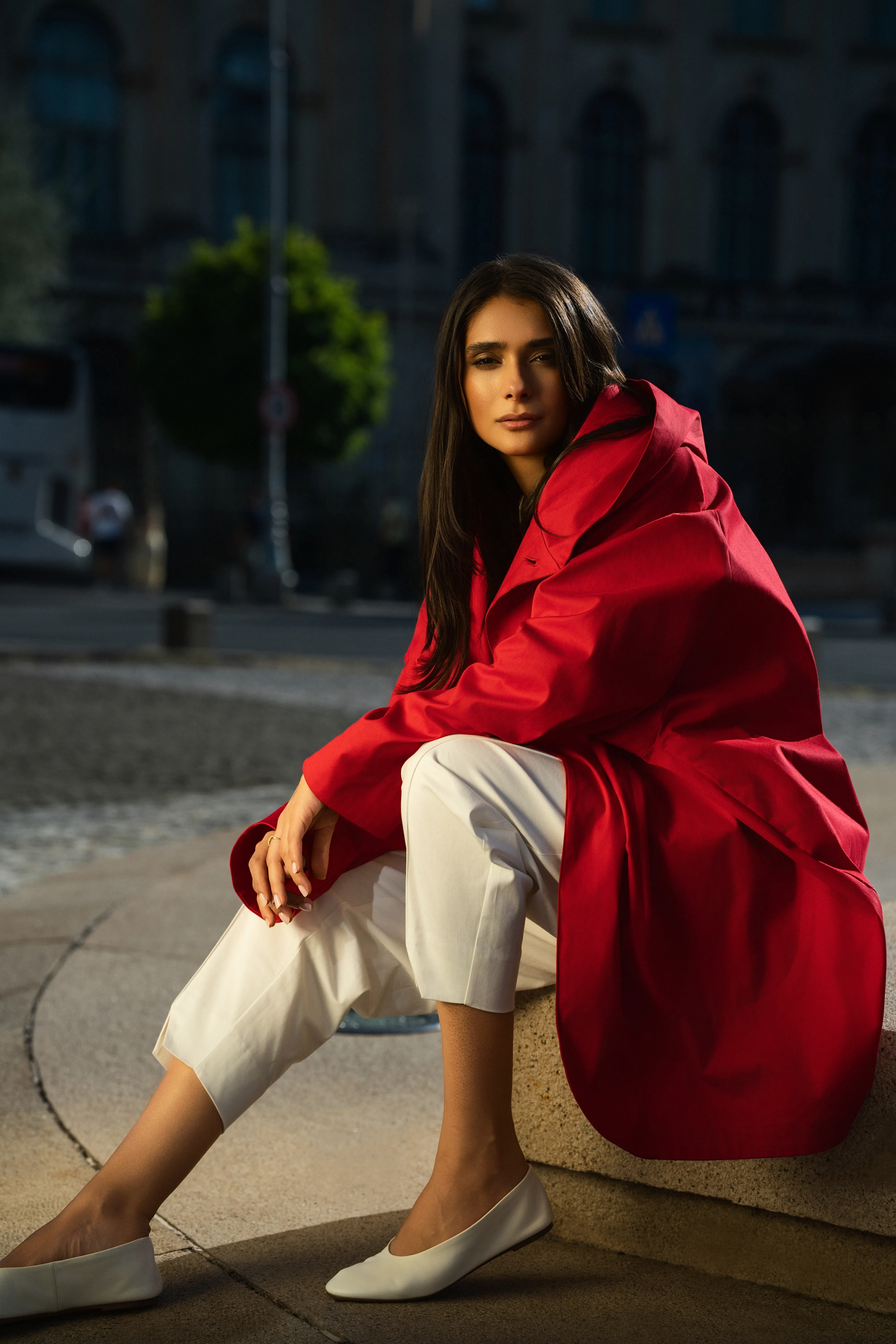 Model in the red hooded coat seated on stone steps in daylight, white trousers