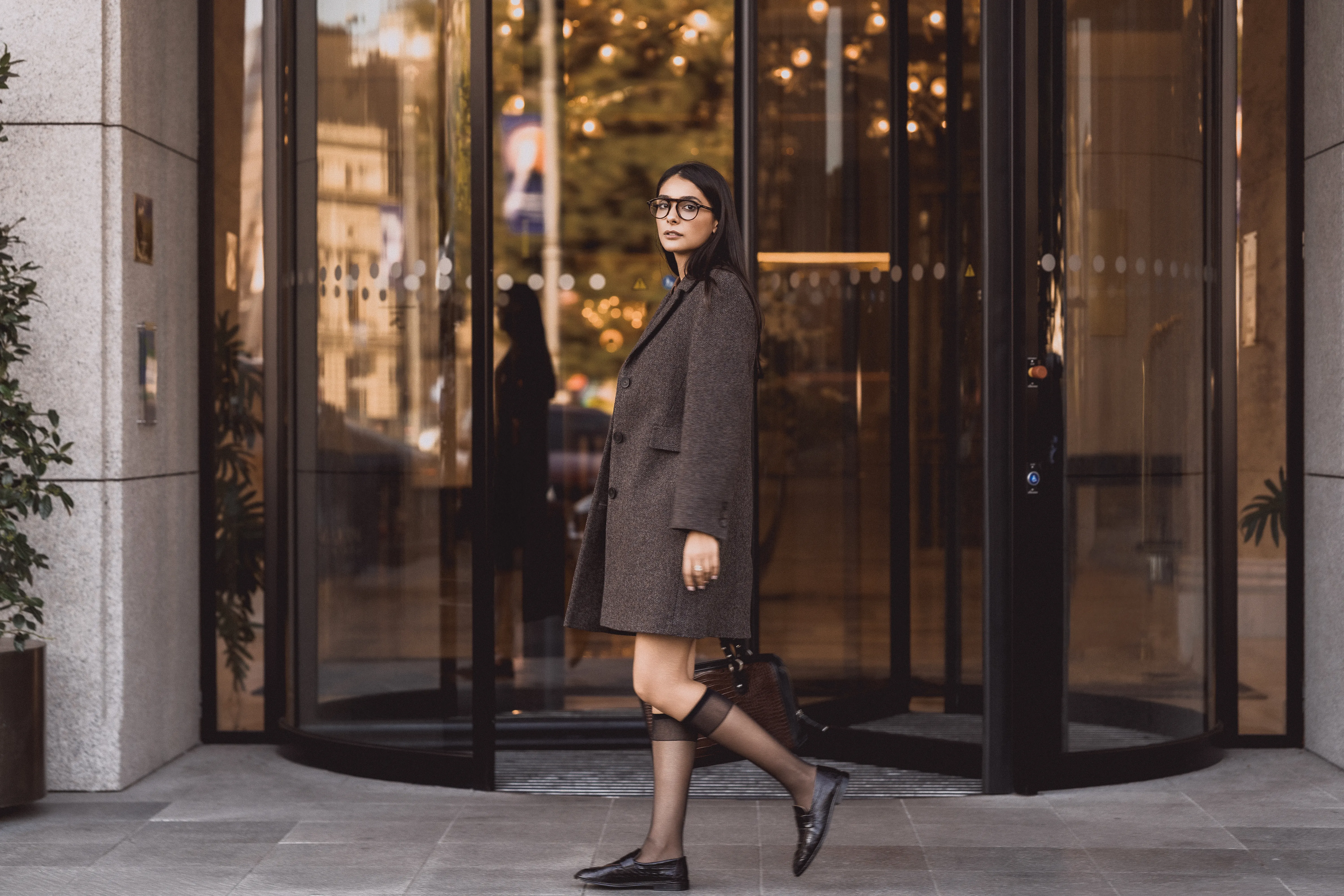 Model in a grey-brown coat walking past a glass revolving door at dusk, reflections