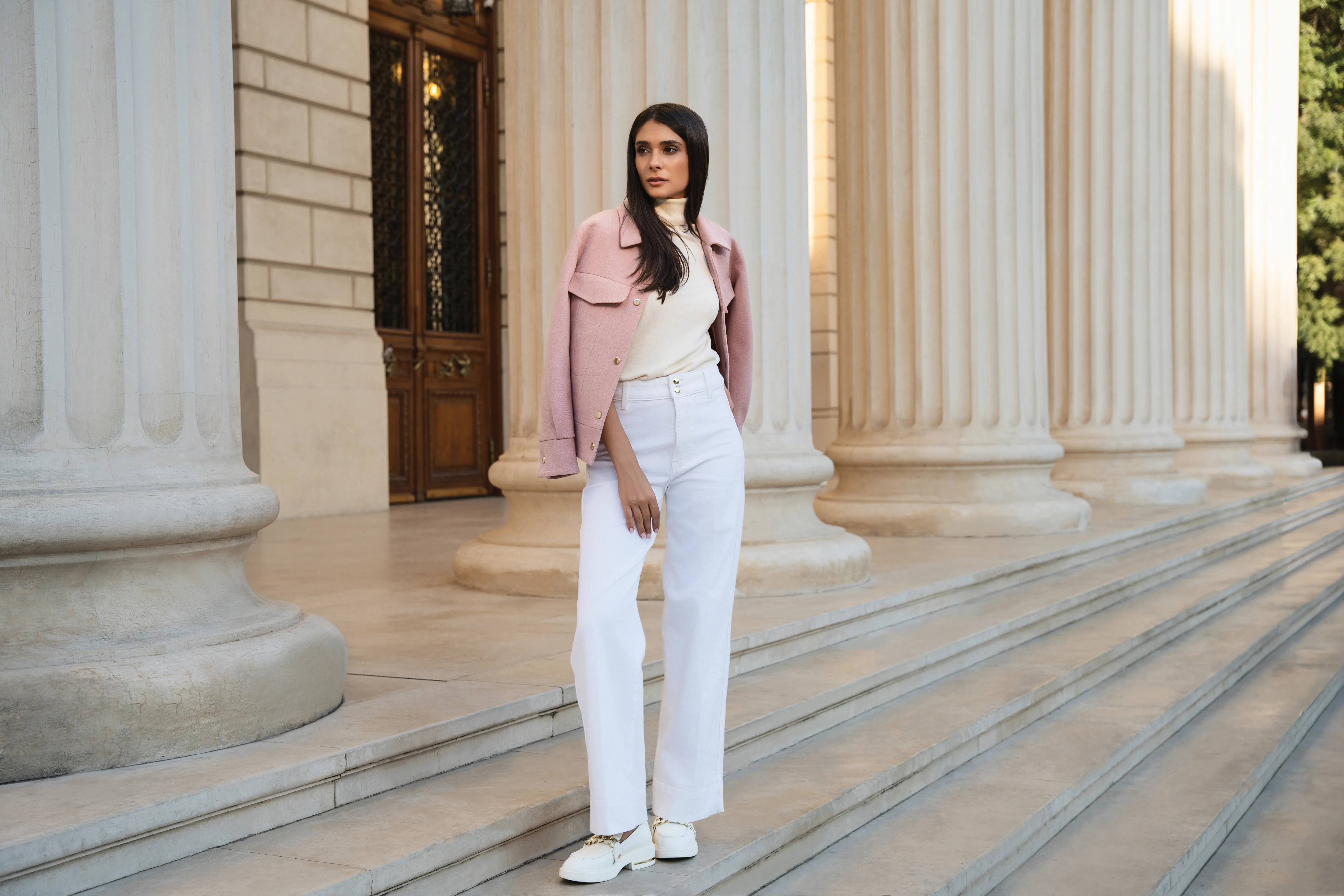 Model in a pink jacket and white trousers walking past a colonnade in soft daylight