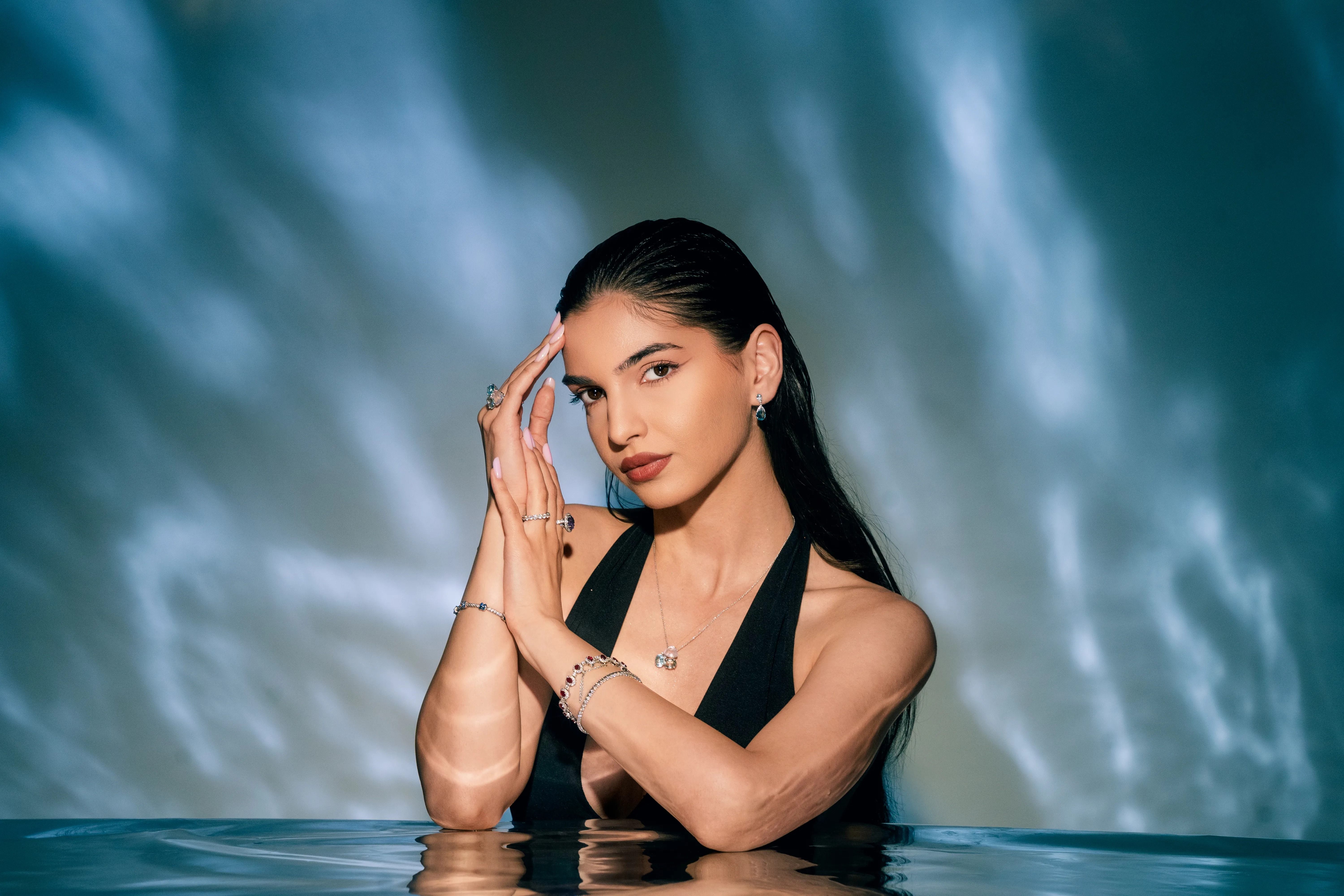 A woman submerged to the shoulders in teal water, jewelry catching dramatic light, hands clasped near her face