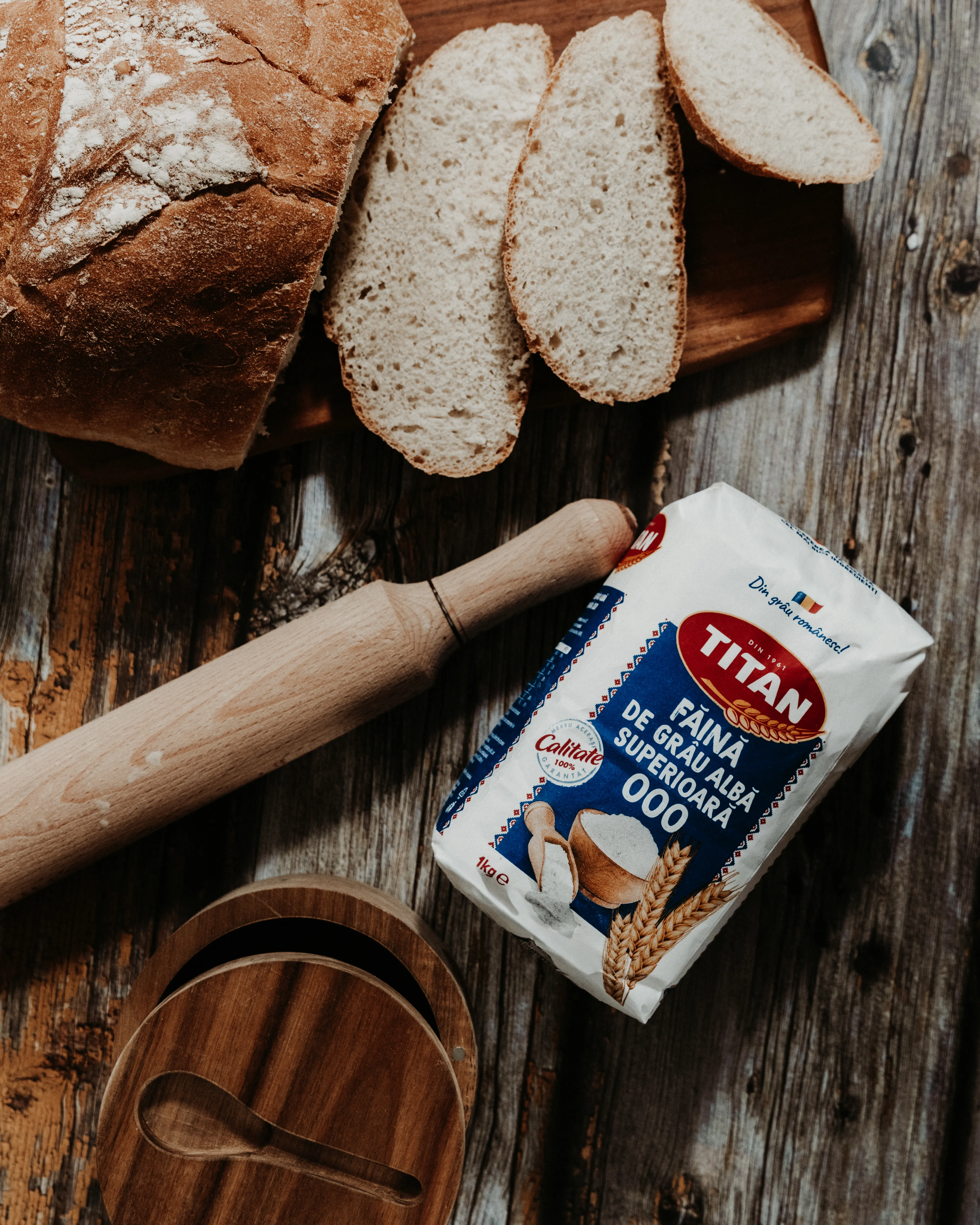 A loaf of bread, sliced, beside a bag of flour on a rustic wooden table