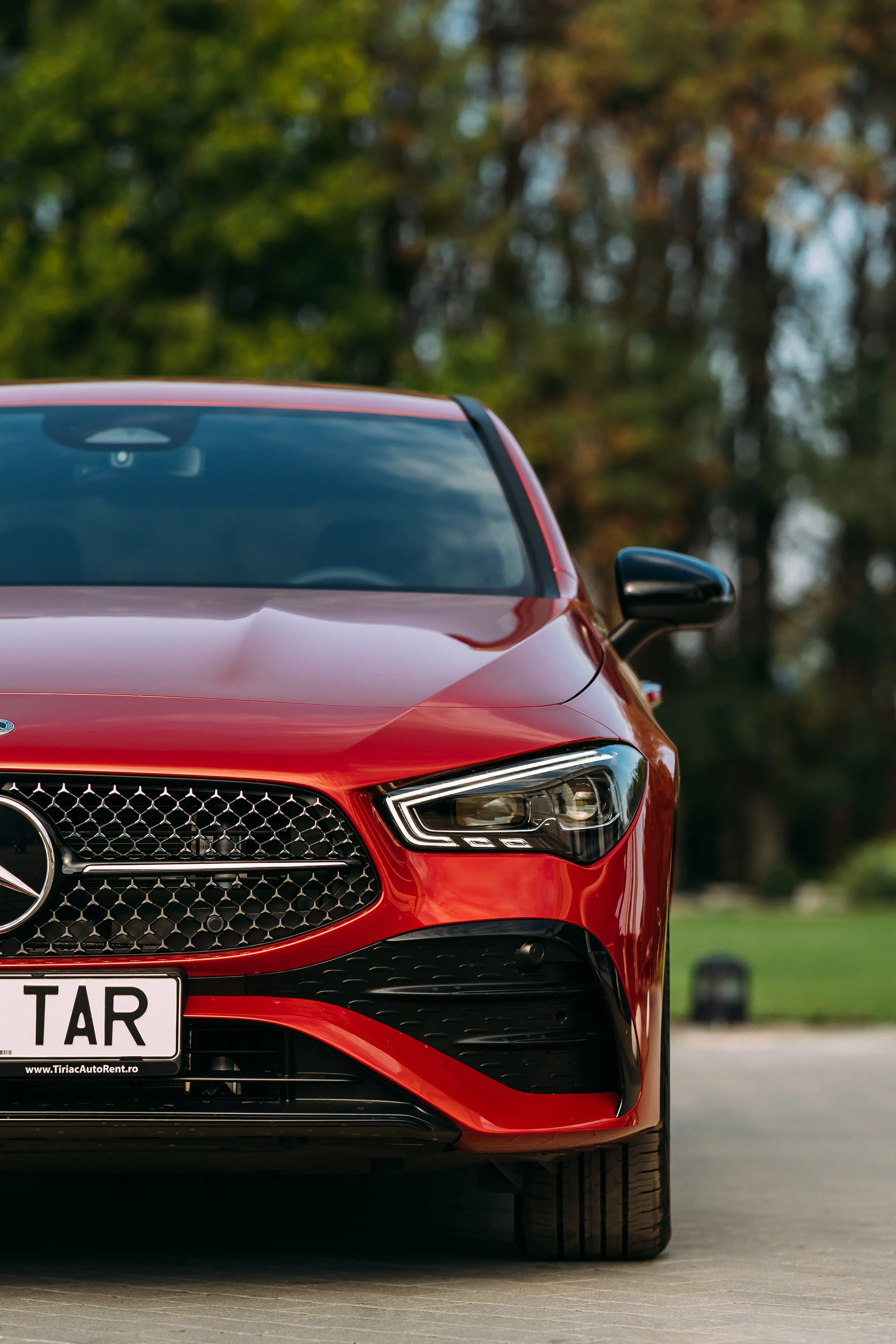 Tight vertical crop of the CLA's front grille and headlight, trees blurred behind
