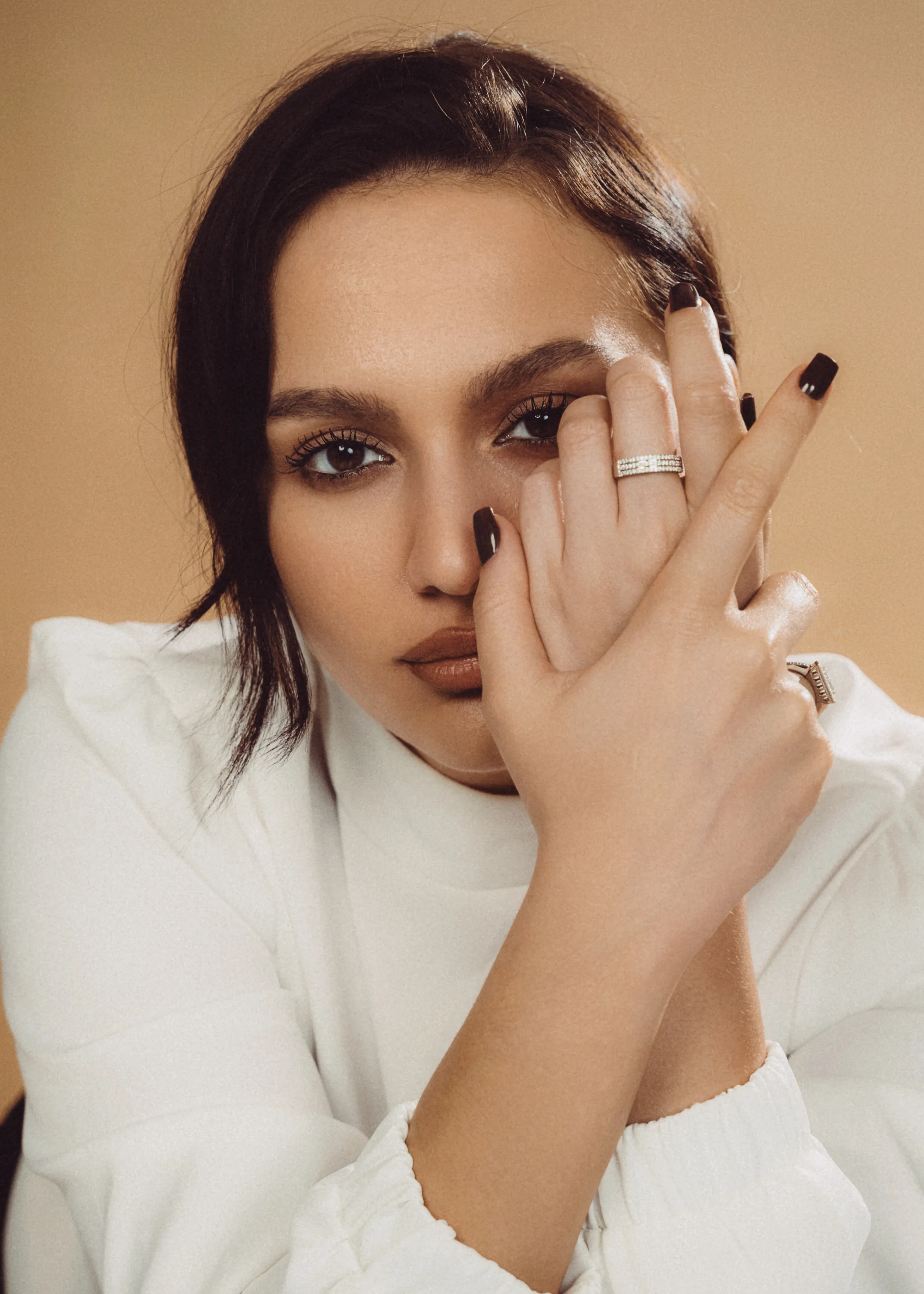 A woman in a white blazer shields half her face with a be-ringed hand, dark manicure, warm backdrop