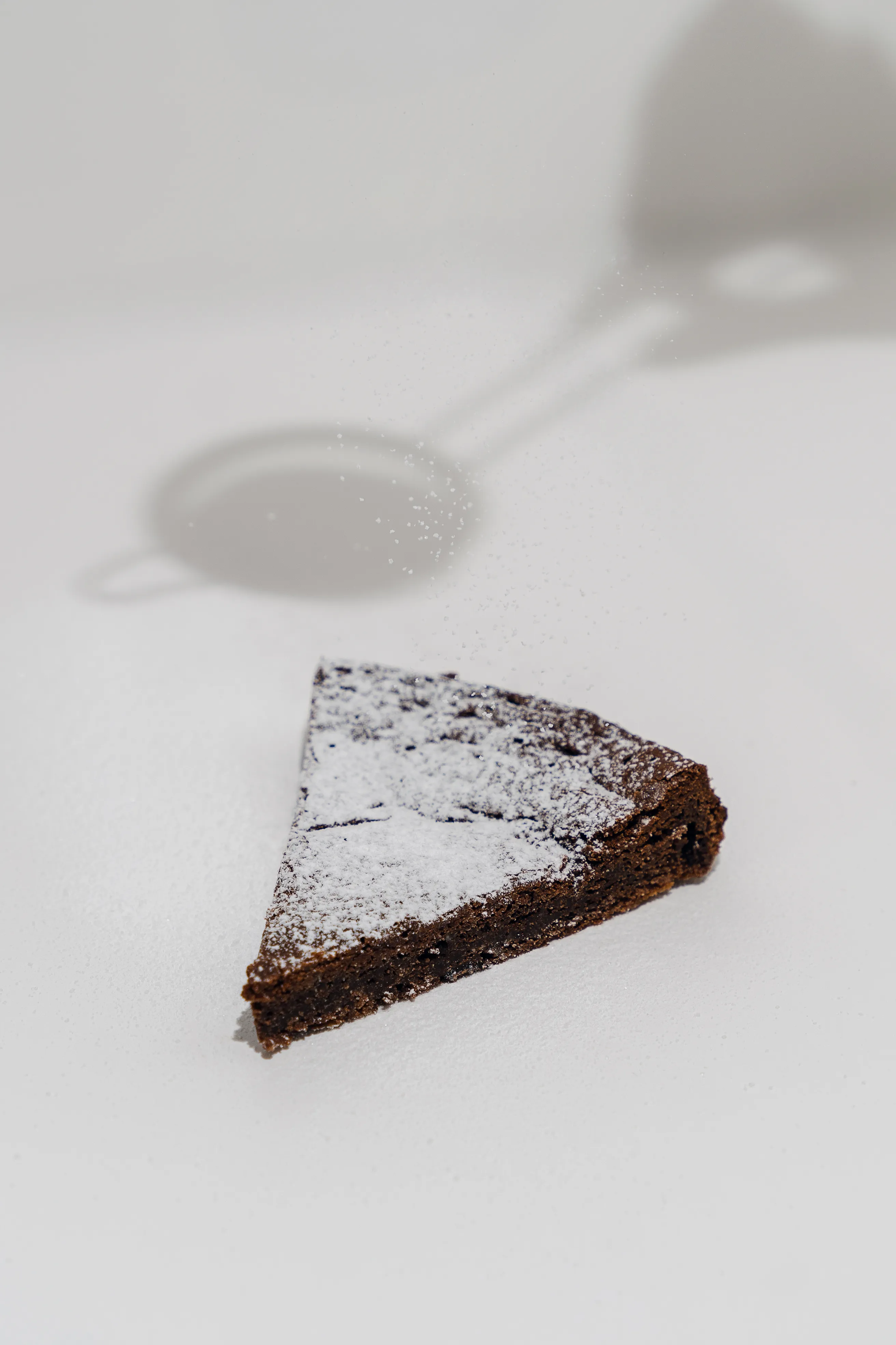 A slice of chocolate cake dusted with powdered sugar mid-fall from a sieve