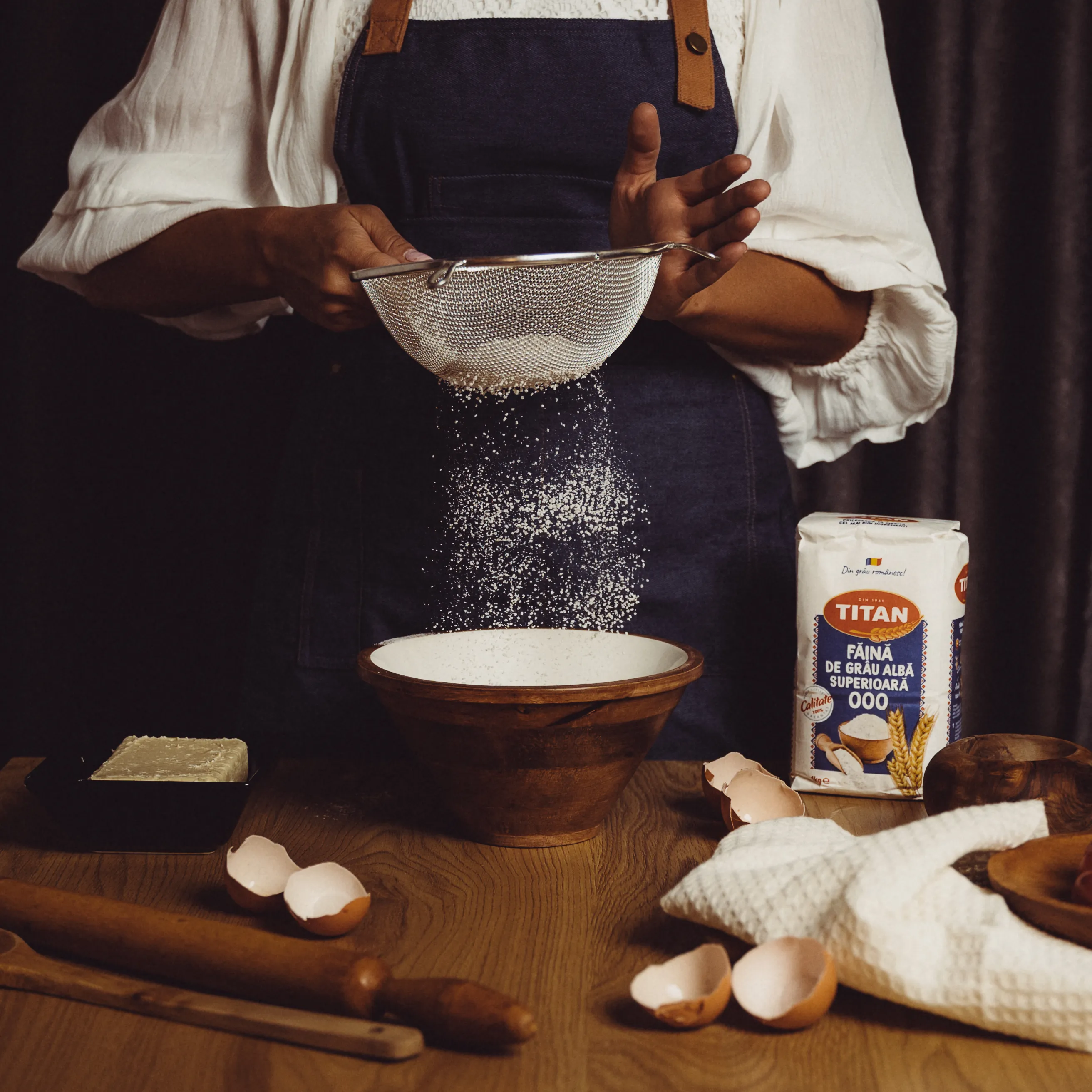 Flour being sifted over a wooden table in dramatic side light, eggshells scattered nearby