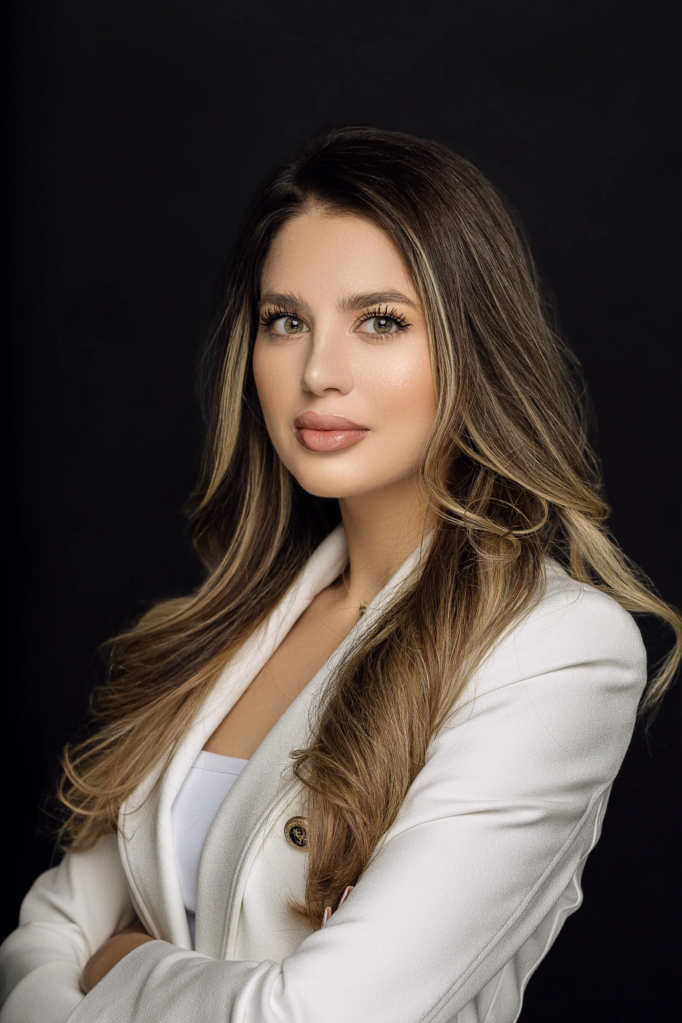 Woman with long hair in a white blazer, arms crossed, studio portrait against a black background