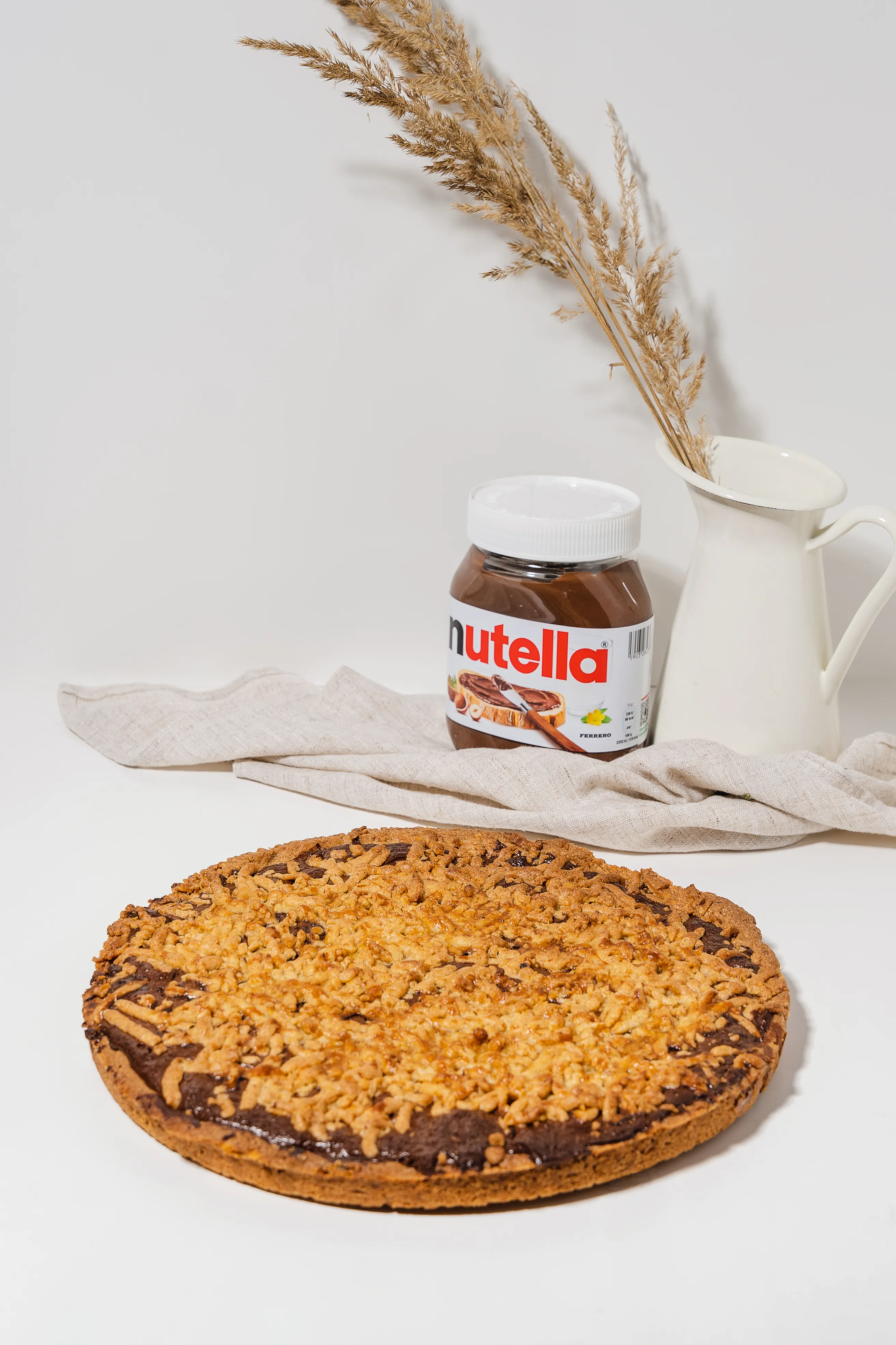 A Nutella crumble cake beside the jar it's made with, dried grasses in a jug behind it