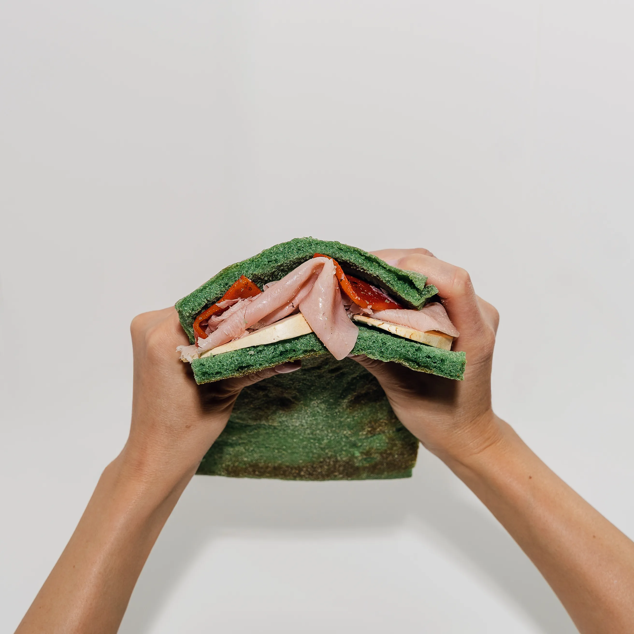 Two hands pulling apart a green herb-bread sandwich filled with cured meats and roasted peppers