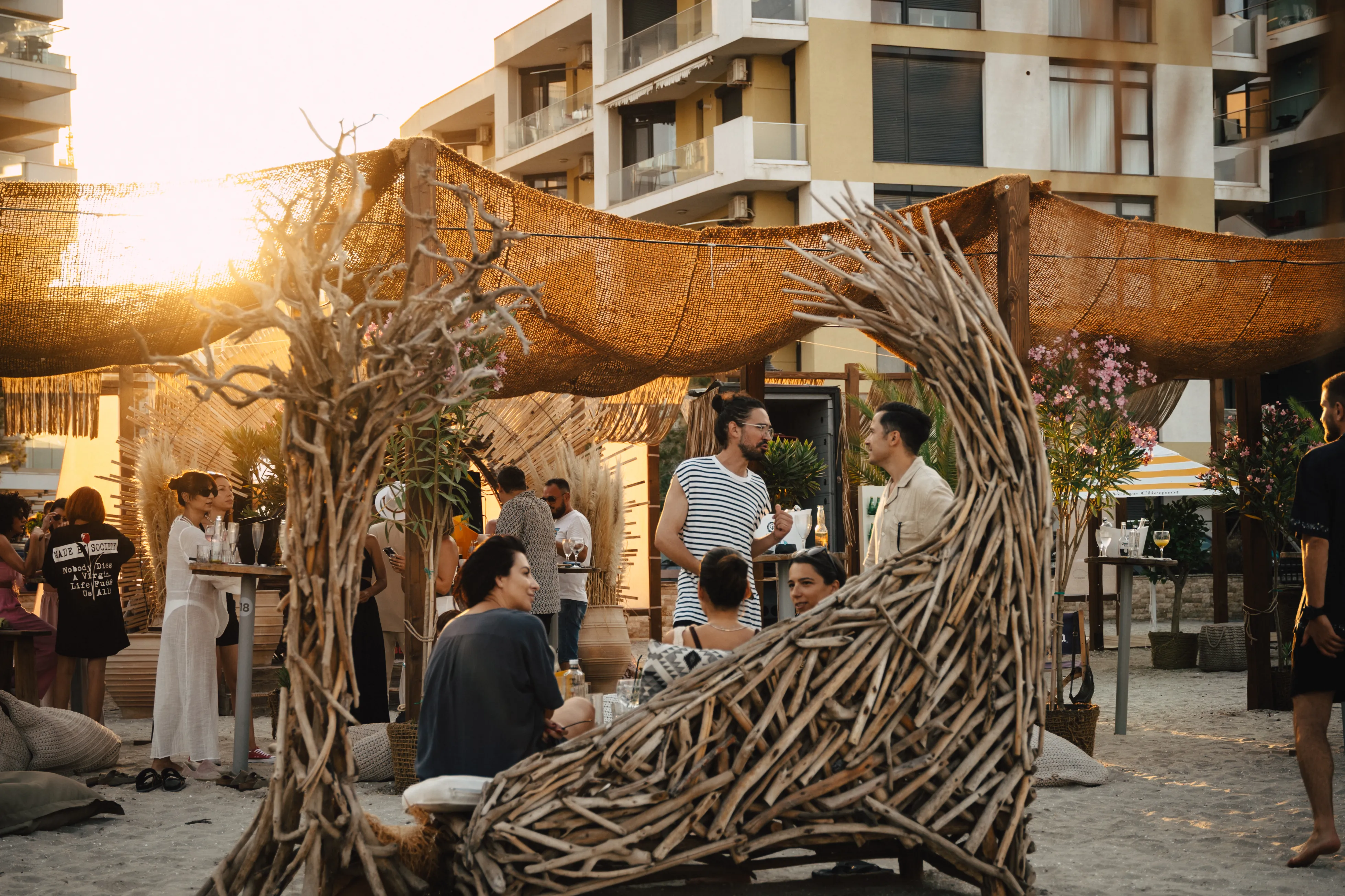 A driftwood pergola frames a golden-hour rooftop gathering, city apartment blocks behind