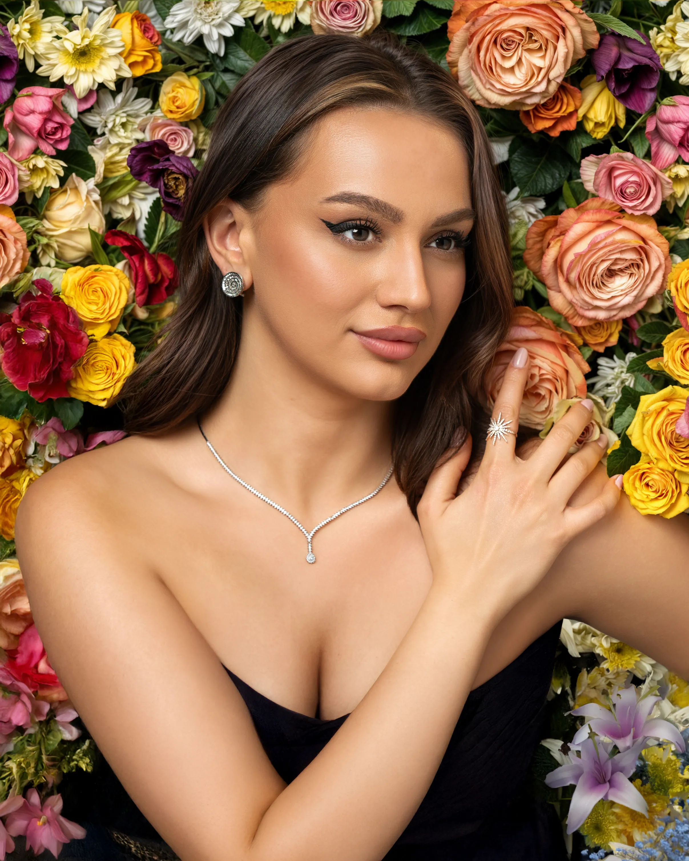 A woman in a black dress poses against a dense wall of colorful roses, wearing a diamond necklace and a starburst ring