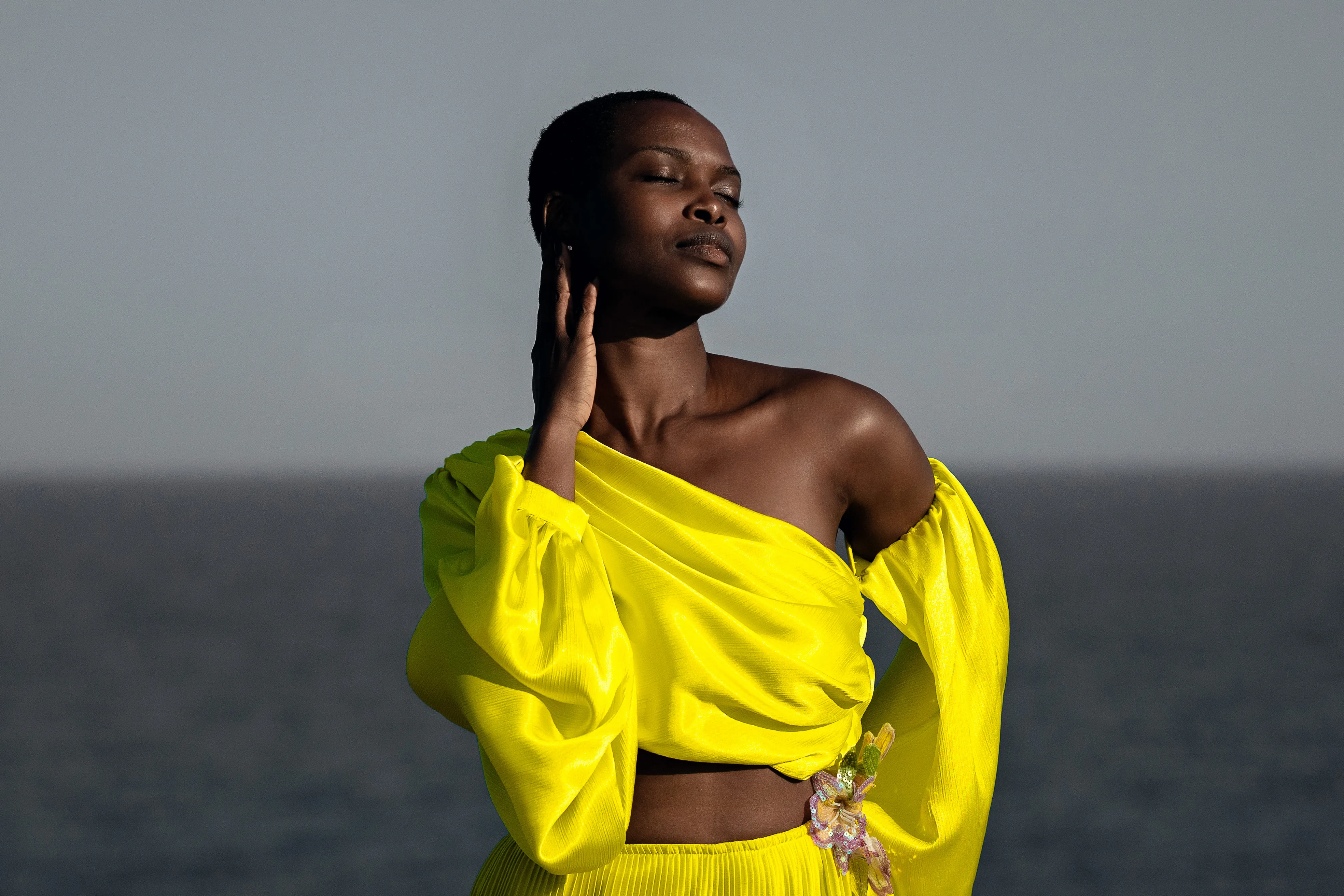 A woman in a bright yellow one-shoulder gown stands before the ocean at golden hour, eyes closed, touching her hair