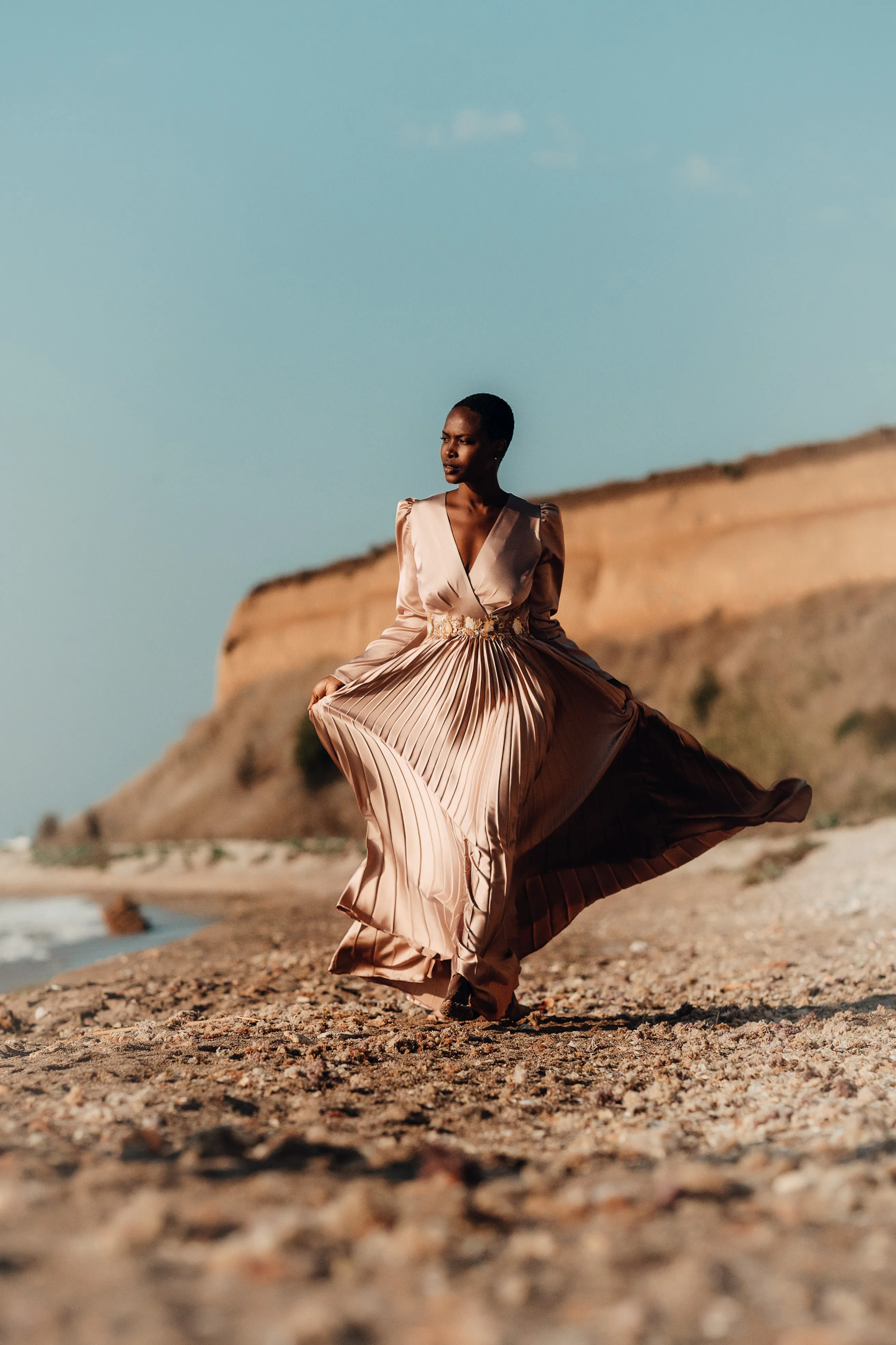 A woman in a flowing champagne gown moves across a beach beneath pale sandstone cliffs