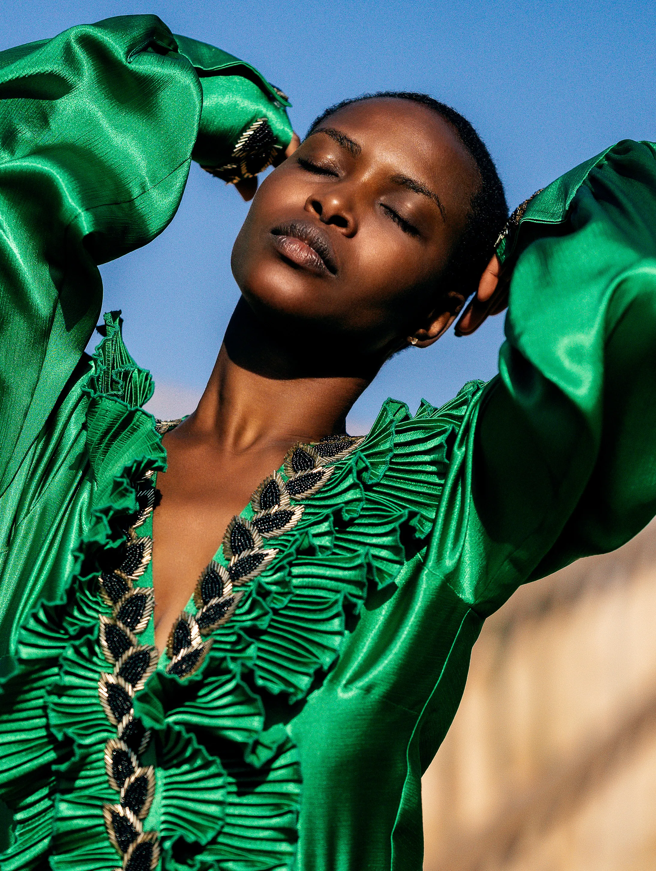 A woman in an emerald pleated gown with beaded trim tilts her face into full sun, eyes closed, against a clear blue sky