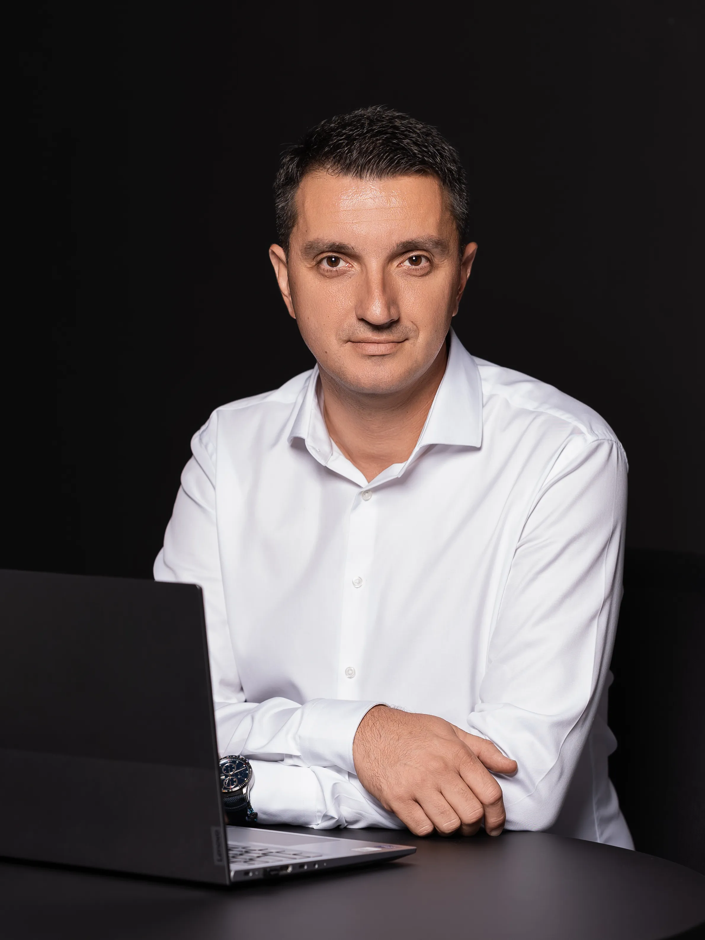 Man in a white dress shirt seated at a laptop, arms crossed, against a dark background
