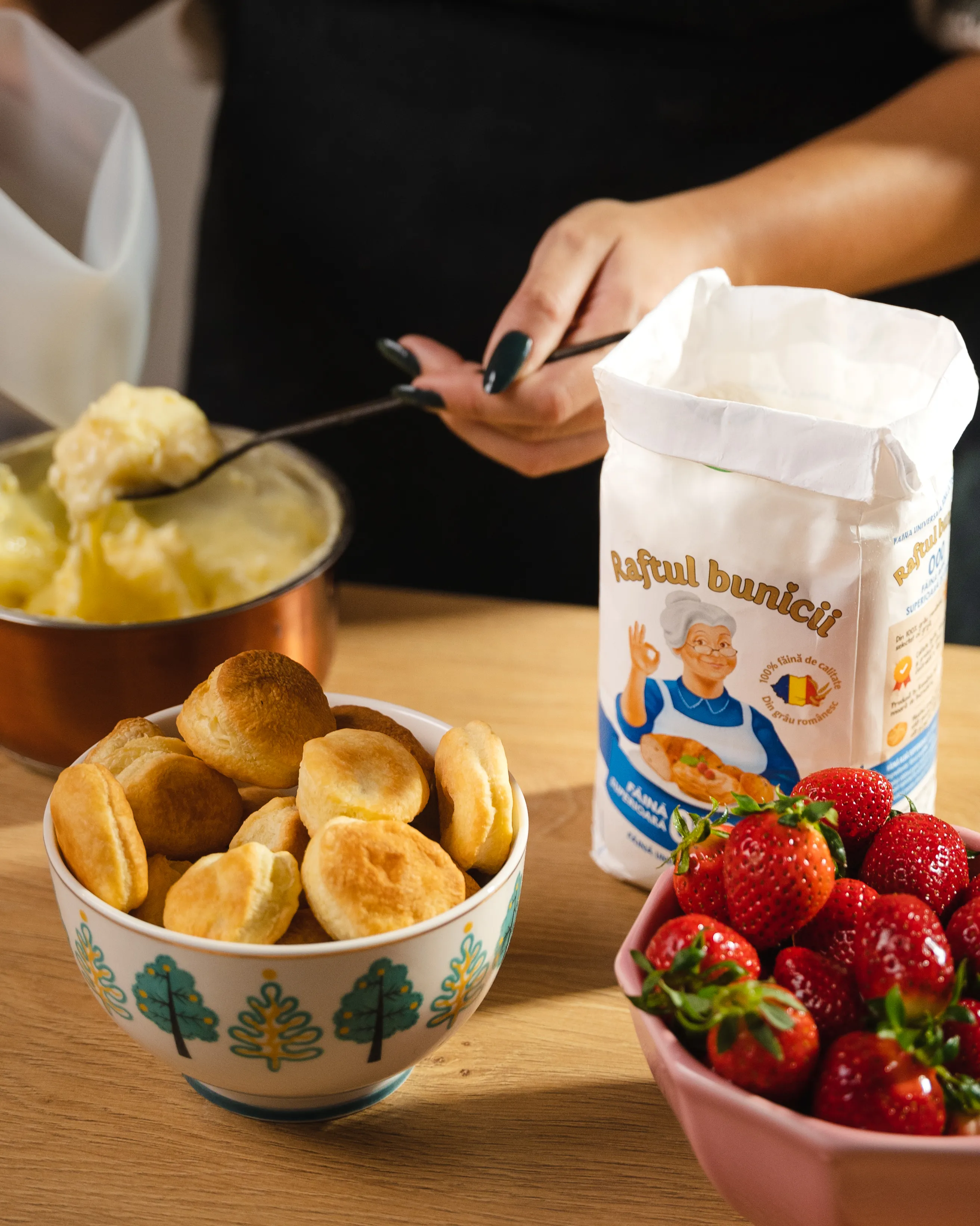 Hands pouring Raftul Bunicii flour over bowls of dough and strawberries