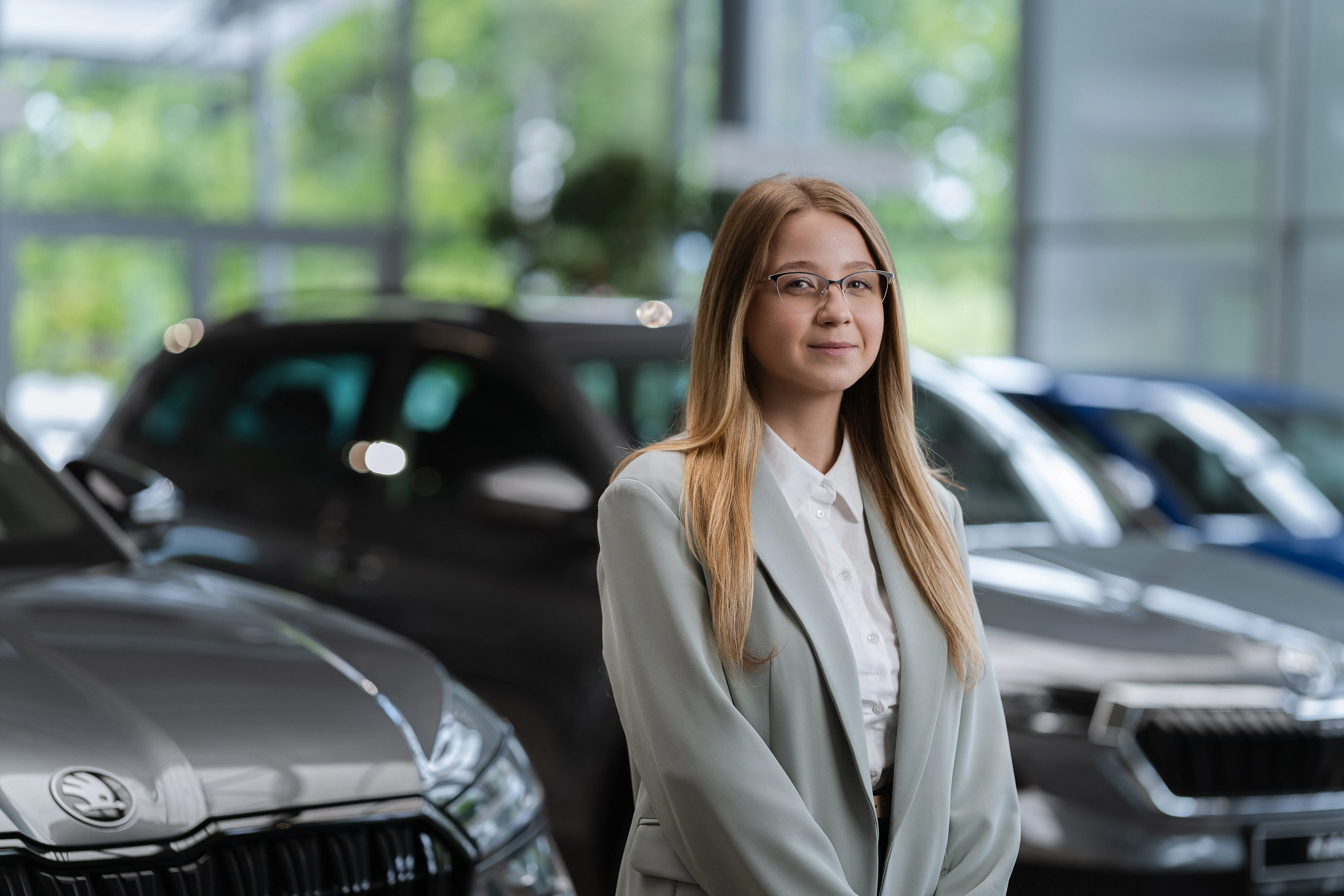 Portrait of an advisor in glasses and a light cardigan, hands clasped, showroom cars behind her