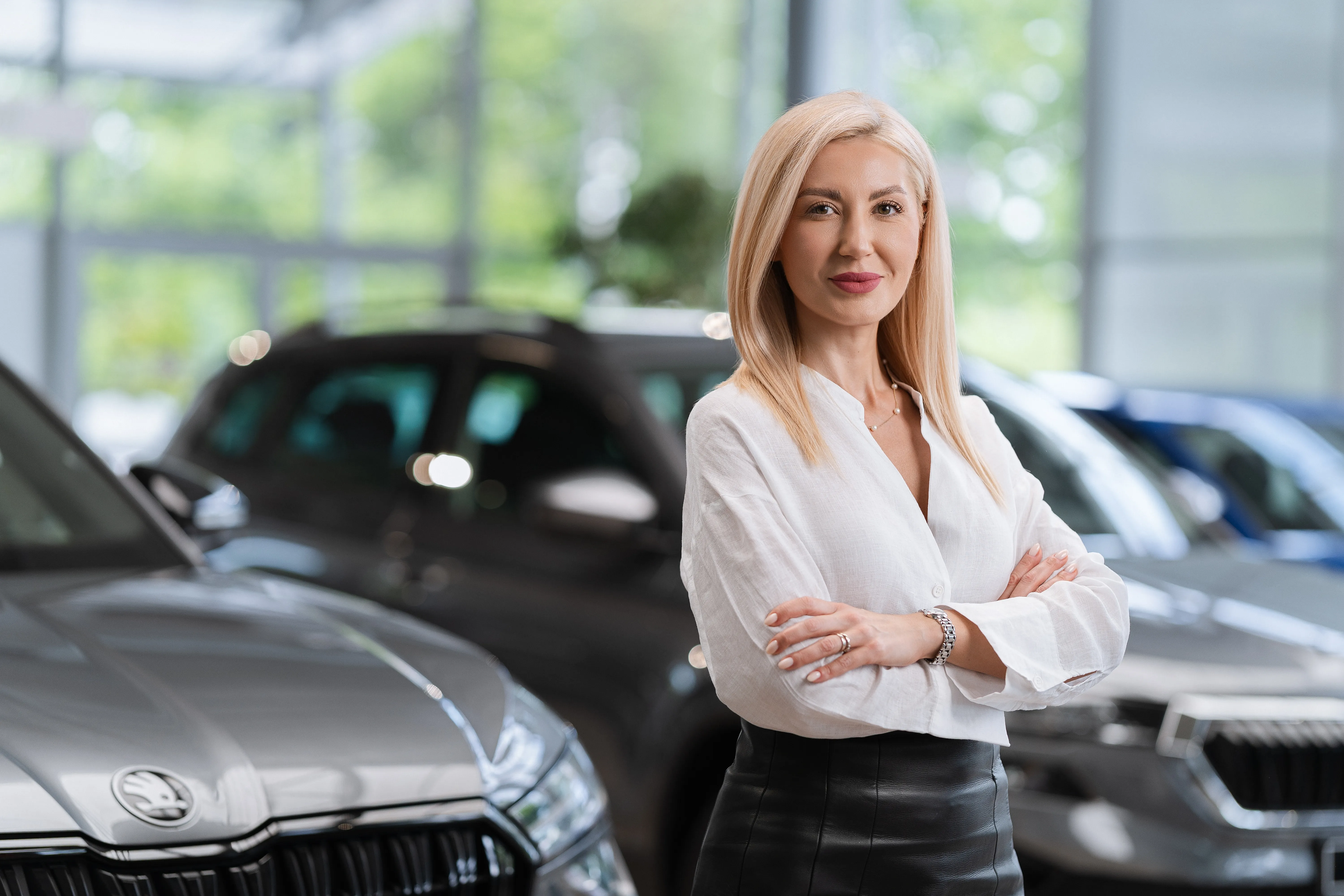 Woman with arms crossed in a car dealership showroom, cars blurred in the background