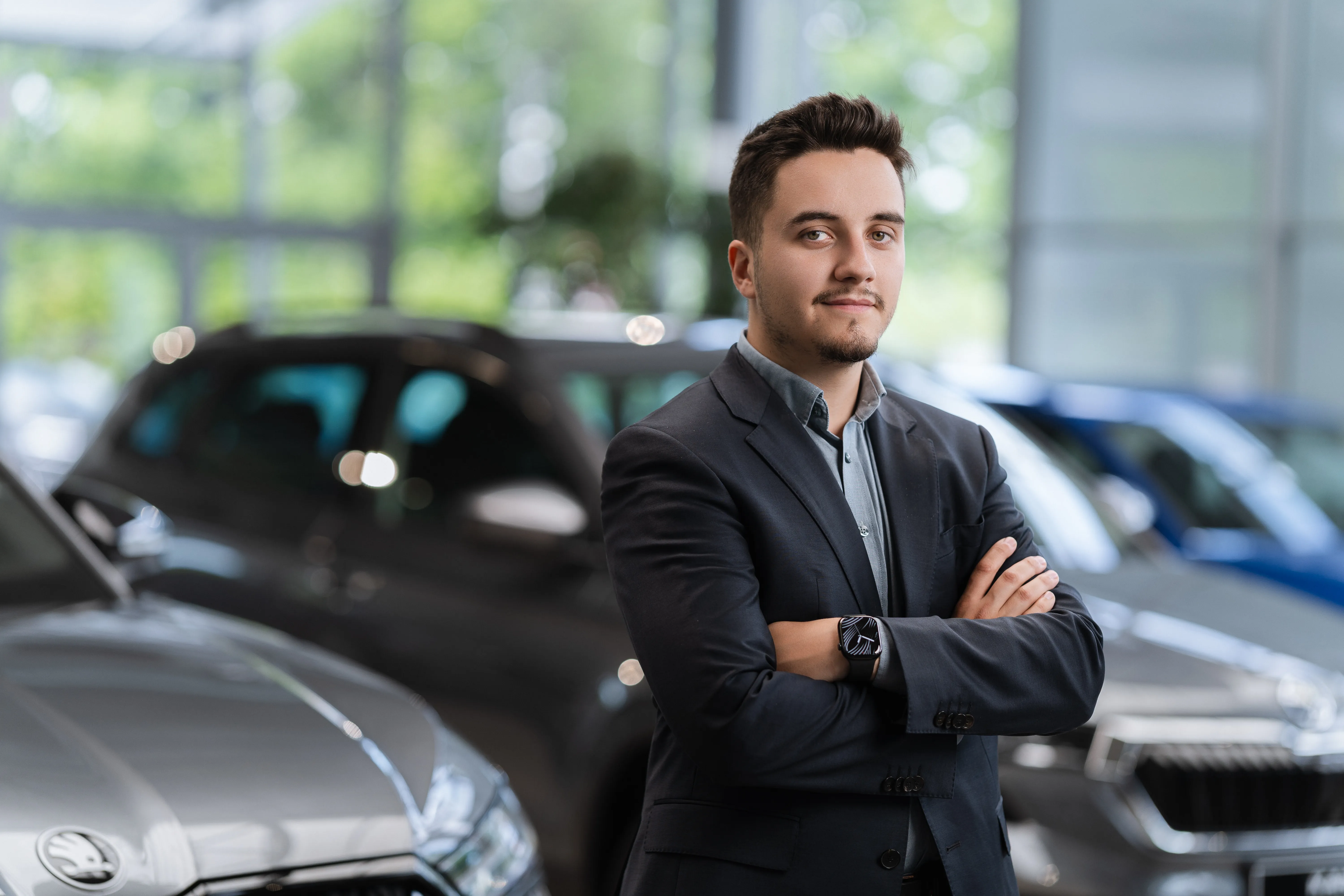 Portrait of a young advisor in a dark suit, arms crossed, showroom cars behind him