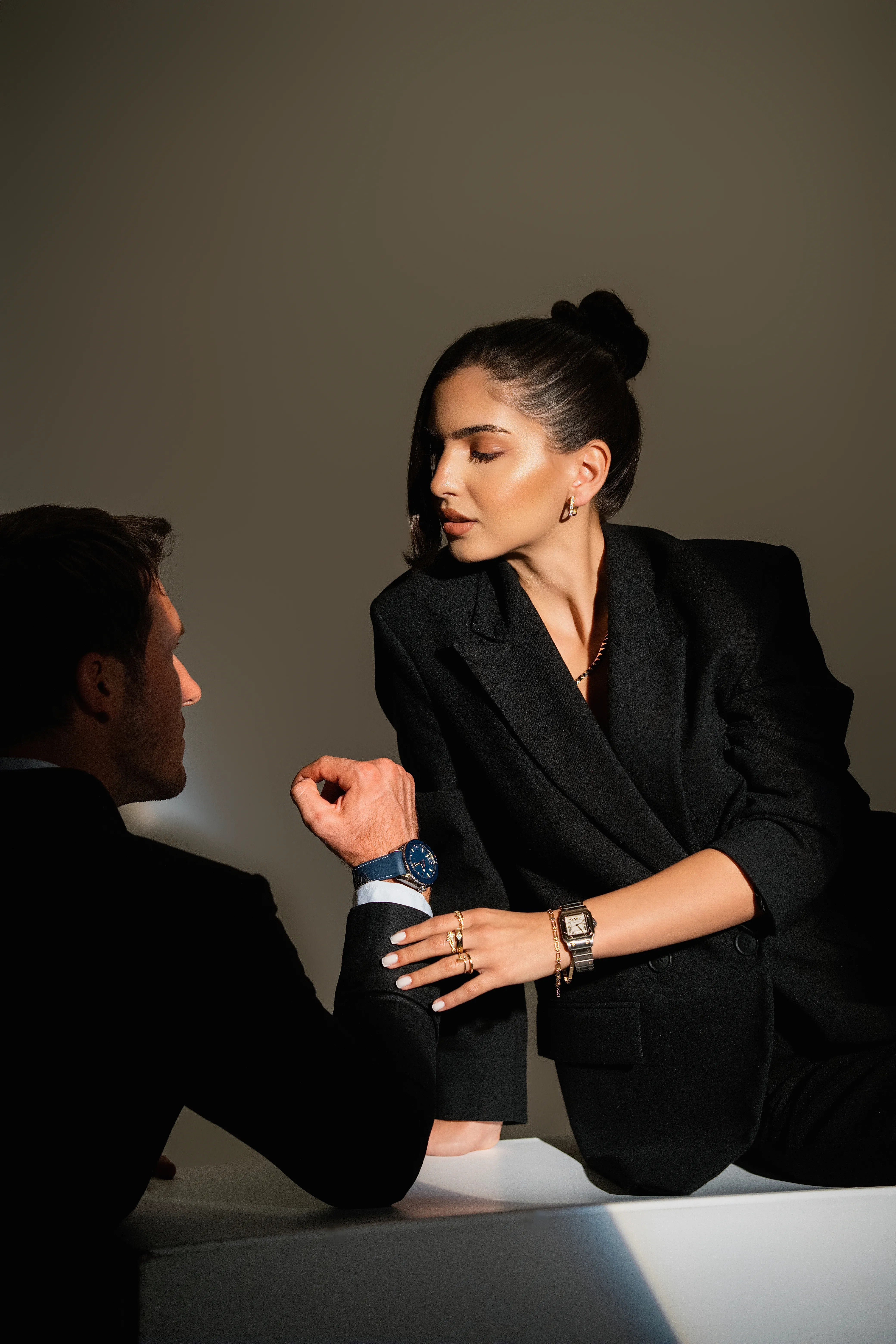 A woman in a black blazer and gold jewelry leans toward a man seated across from her, both wearing watches