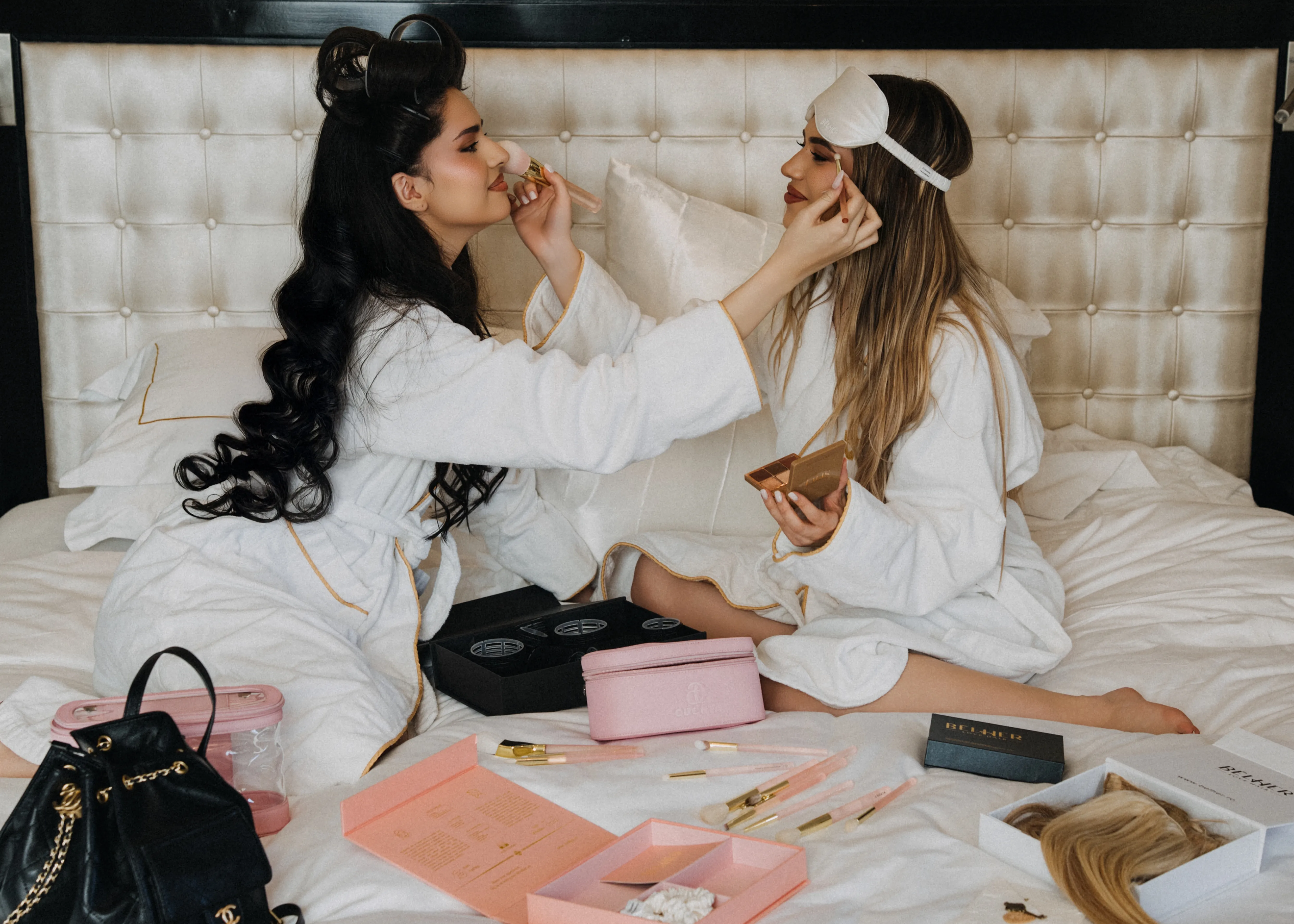Two women in white robes doing each other's makeup on a bed, beauty cases and a wig box scattered around them