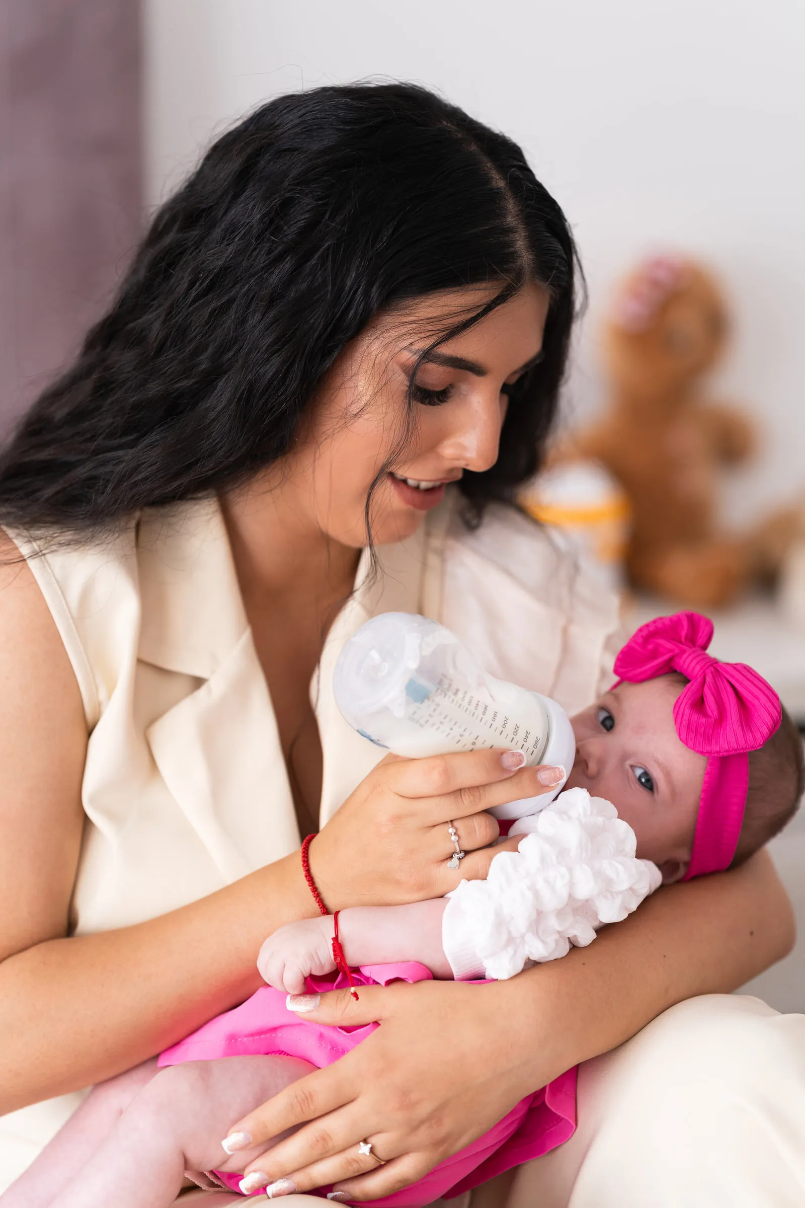 A mother smiles down as she bottle-feeds the baby, who wears a bright pink bow