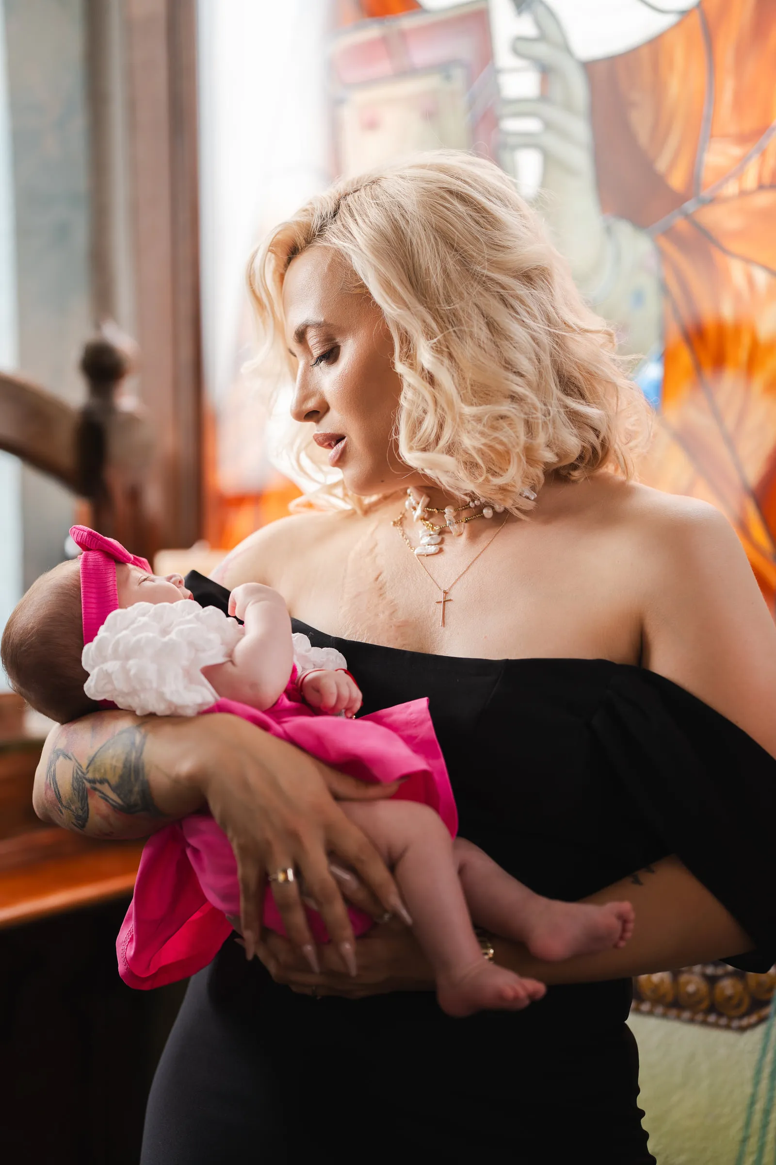 The godmother holds the baby in a pink dress in warm window light, looking down at her