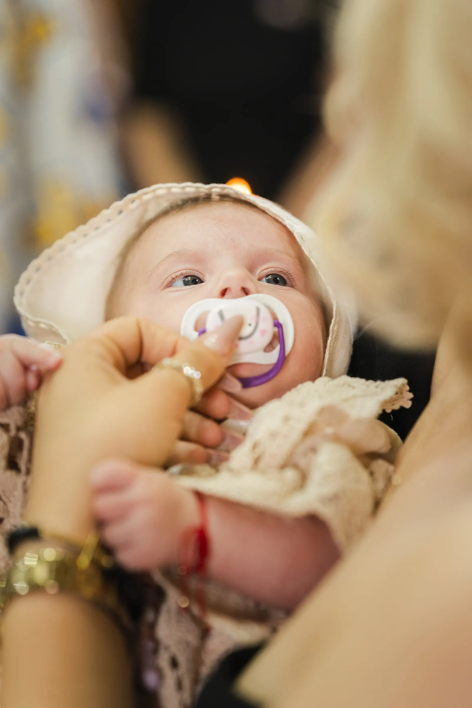 Close view of the baby in a cream lace bonnet, a pacifier in her mouth, held in the church