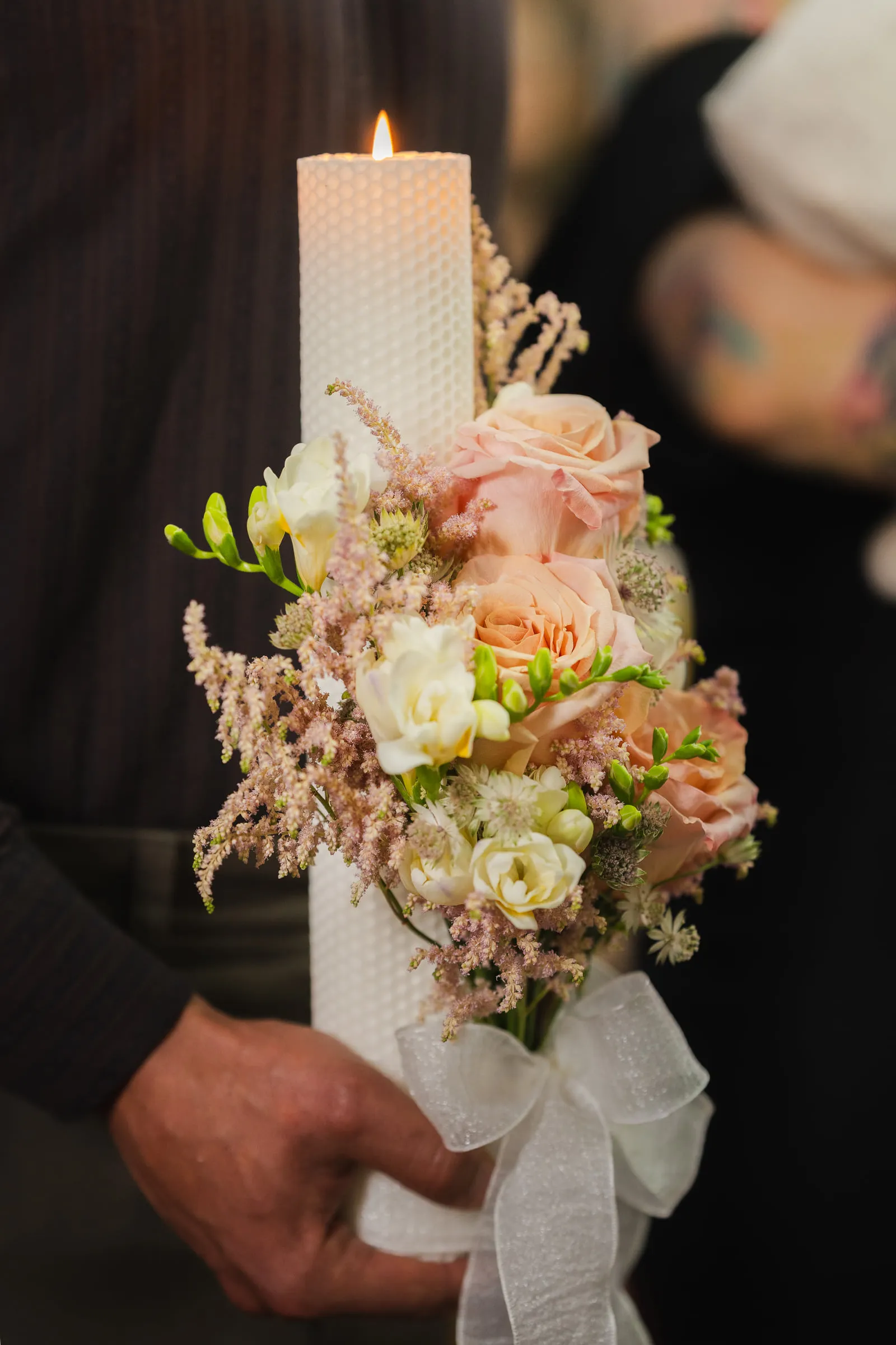 A hand holds the lit baptism candle, wrapped in blush roses and white ribbon