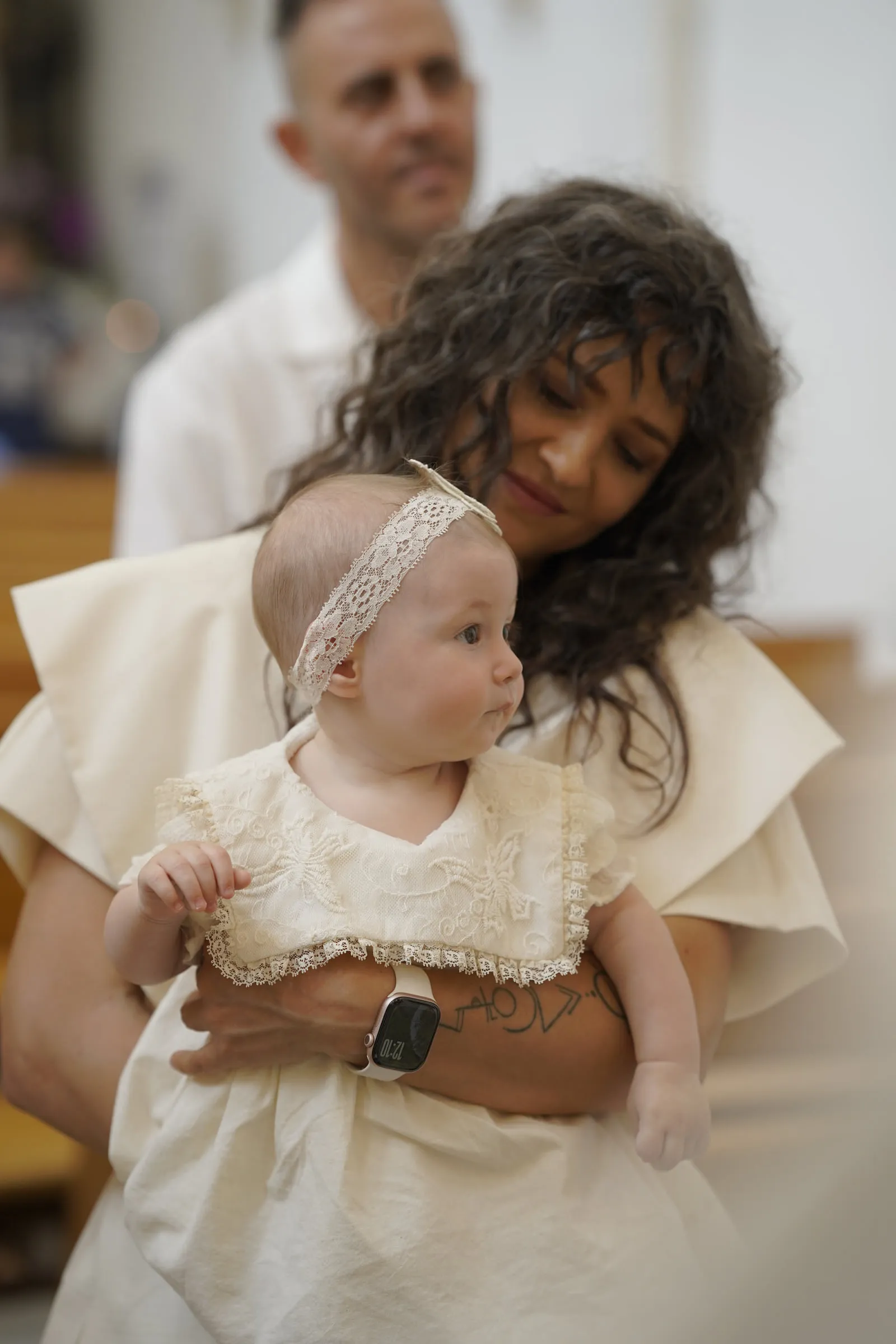 A woman holds the baby, dressed in a lace headband and cream dress, inside the church