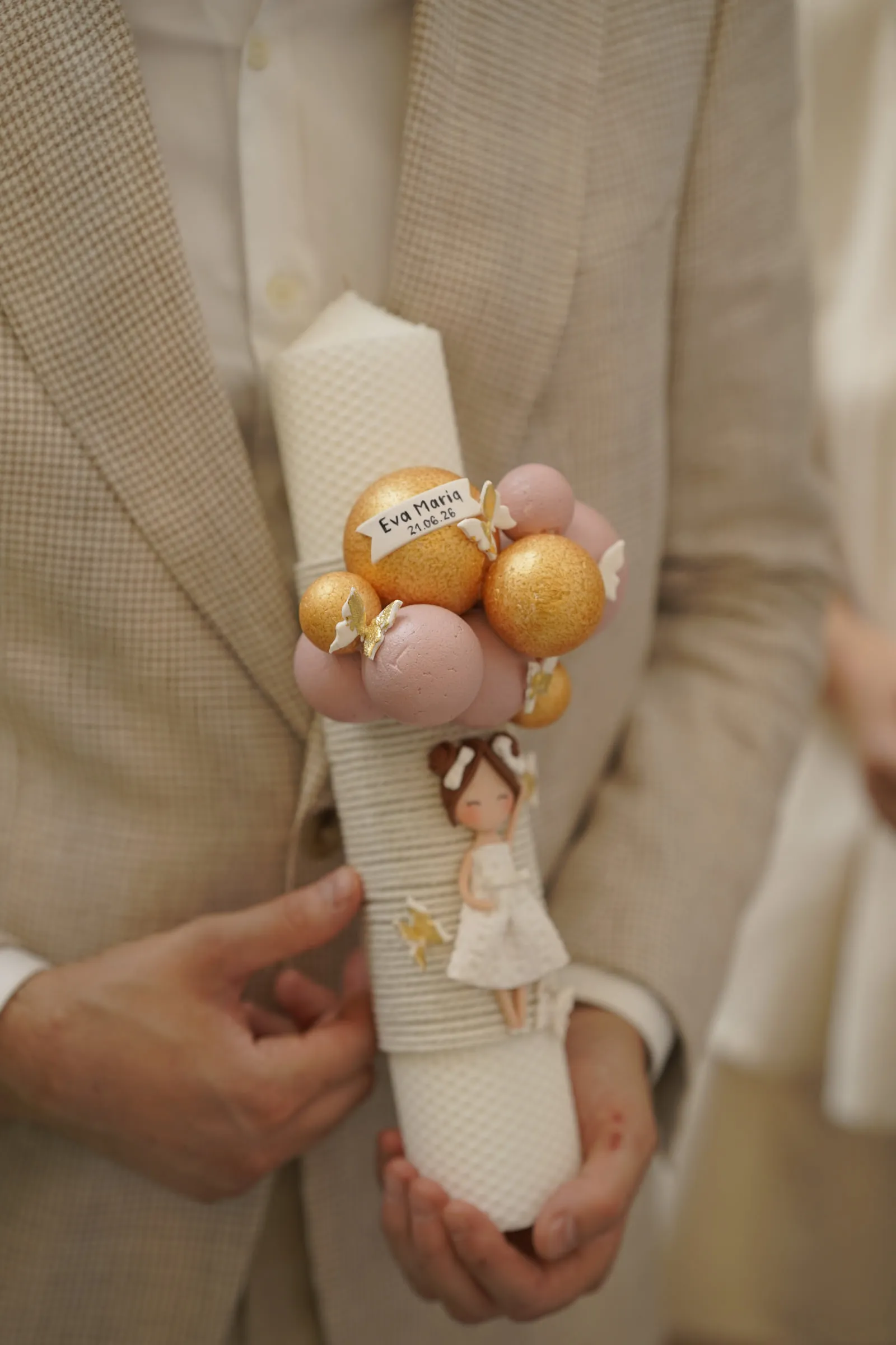 Close detail of hands holding the decorated baptism candle, tied with pink and gold baubles and a name tag reading Eva Maria