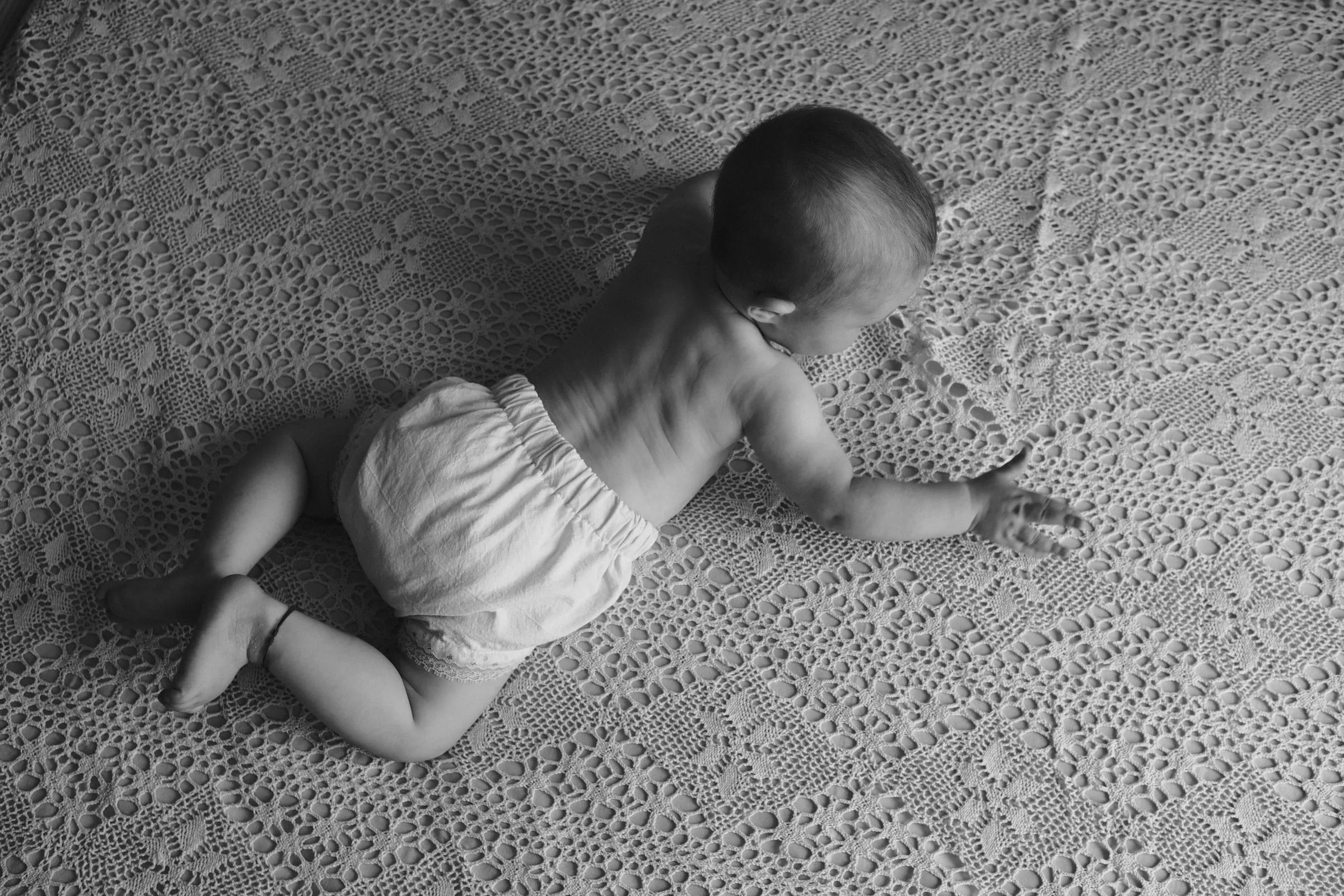 Black and white view from above of the baby crawling across a lace blanket