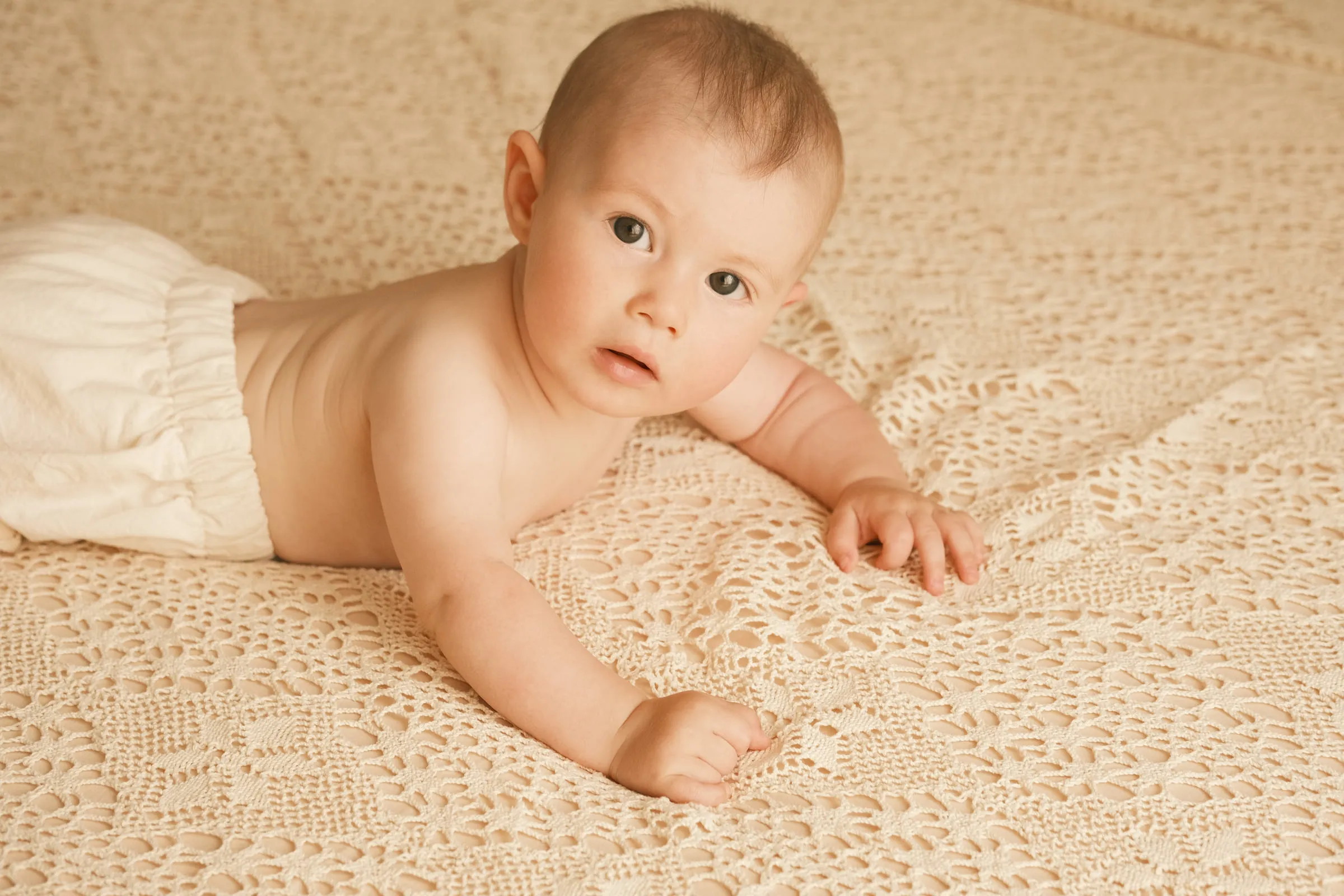 Baby Eva Maria lies on a cream lace blanket, lifting her head to look straight at the camera