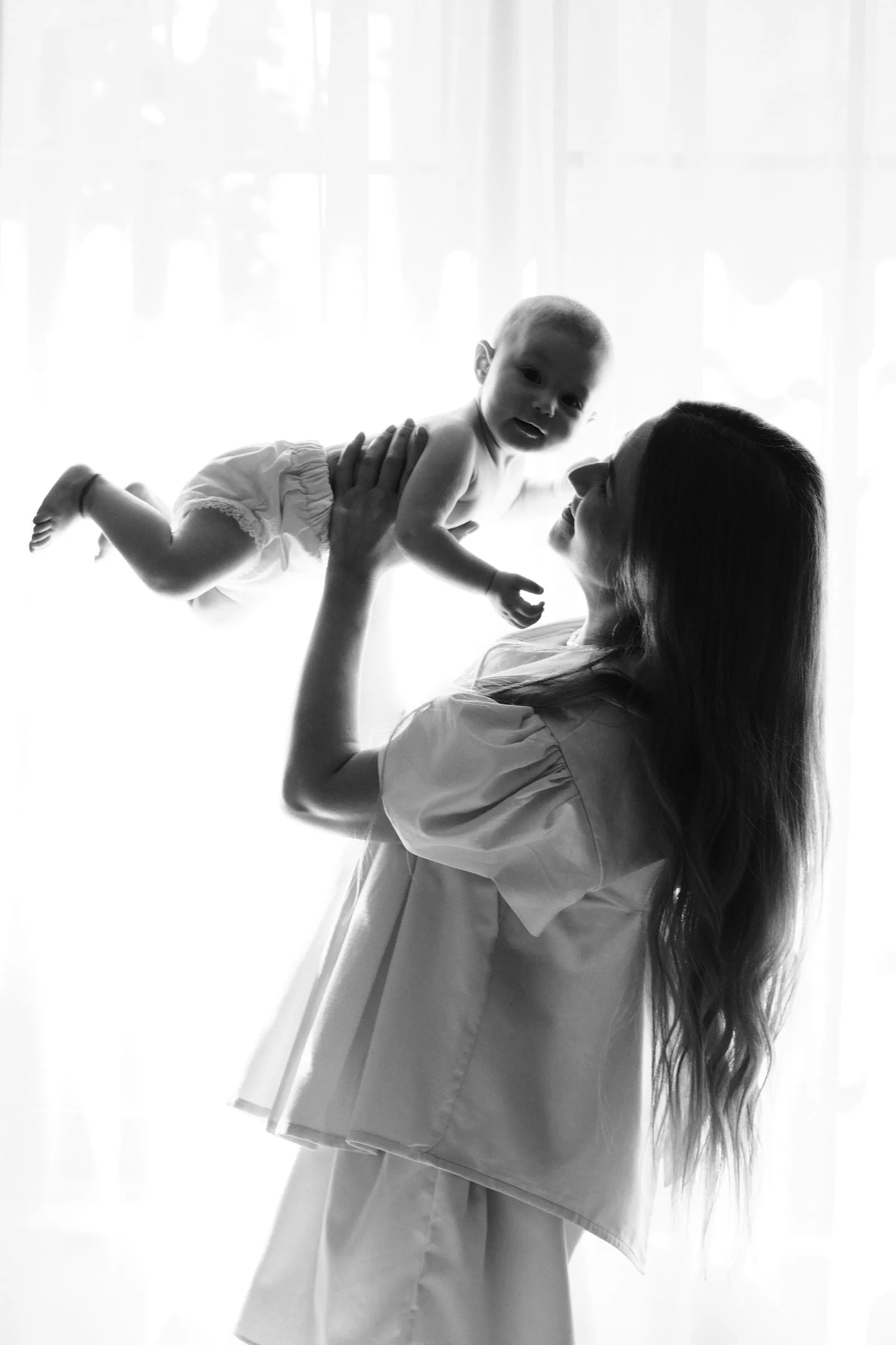 Backlit against a bright window, a mother lifts her baby high above her head