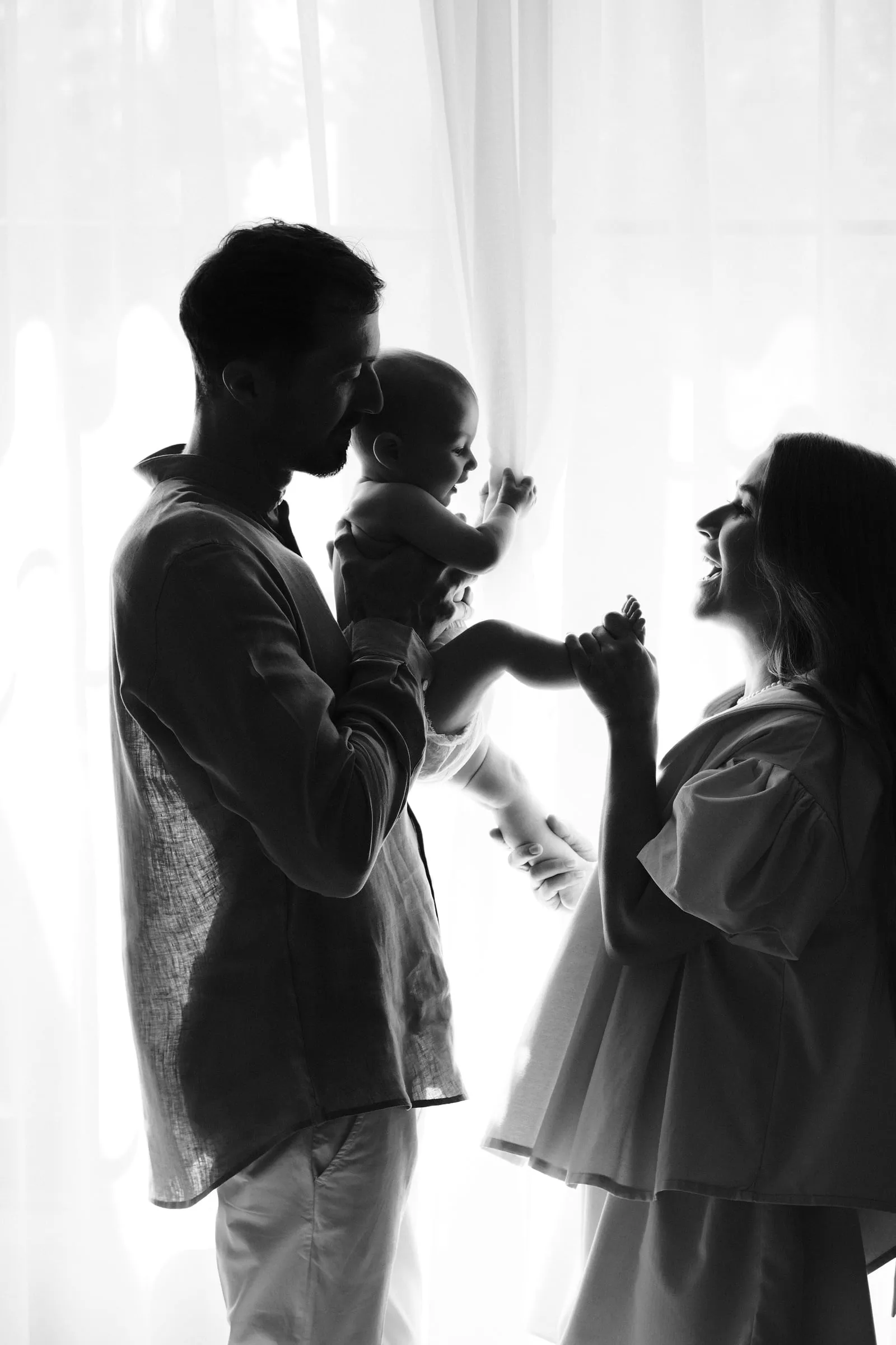 In black and white silhouette, a father kisses the baby he holds while the mother leans in smiling