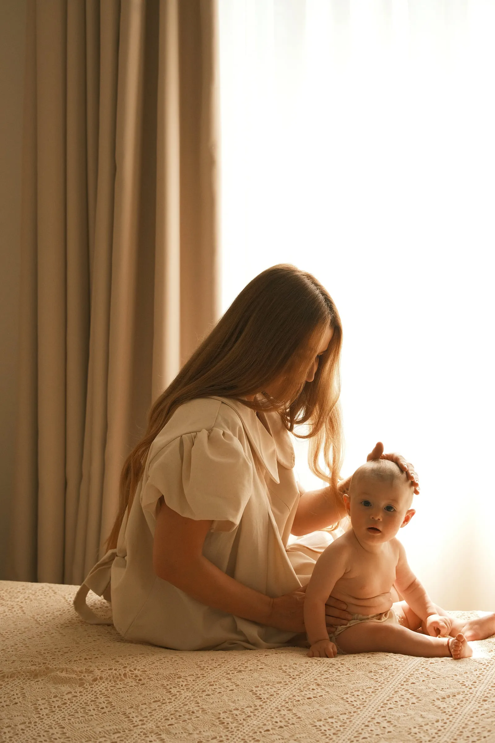 In warm window light, a mother sits on the bed and gently touches her baby's head