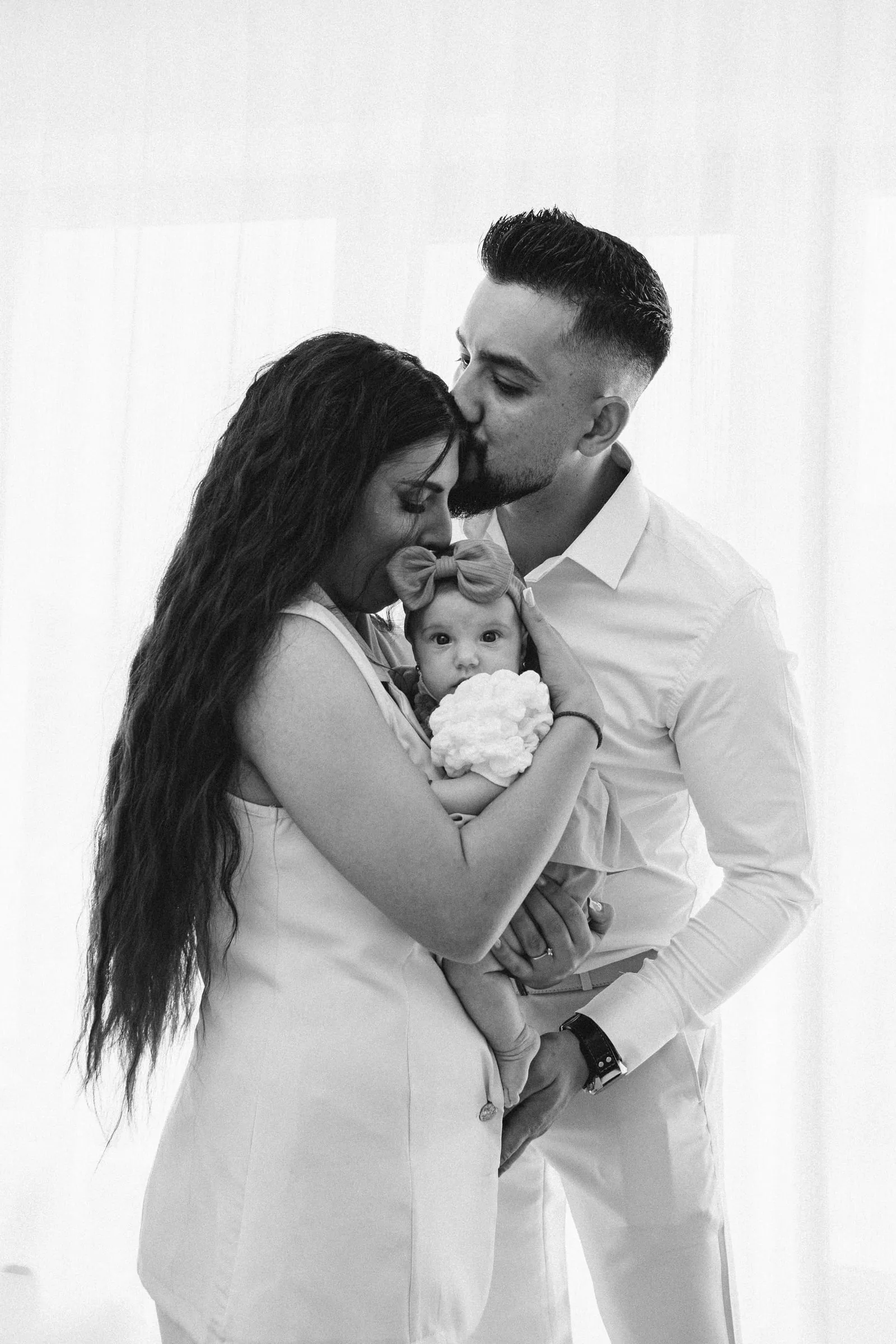 Black and white portrait of the parents embracing, the father kissing the mother's forehead as she holds the baby