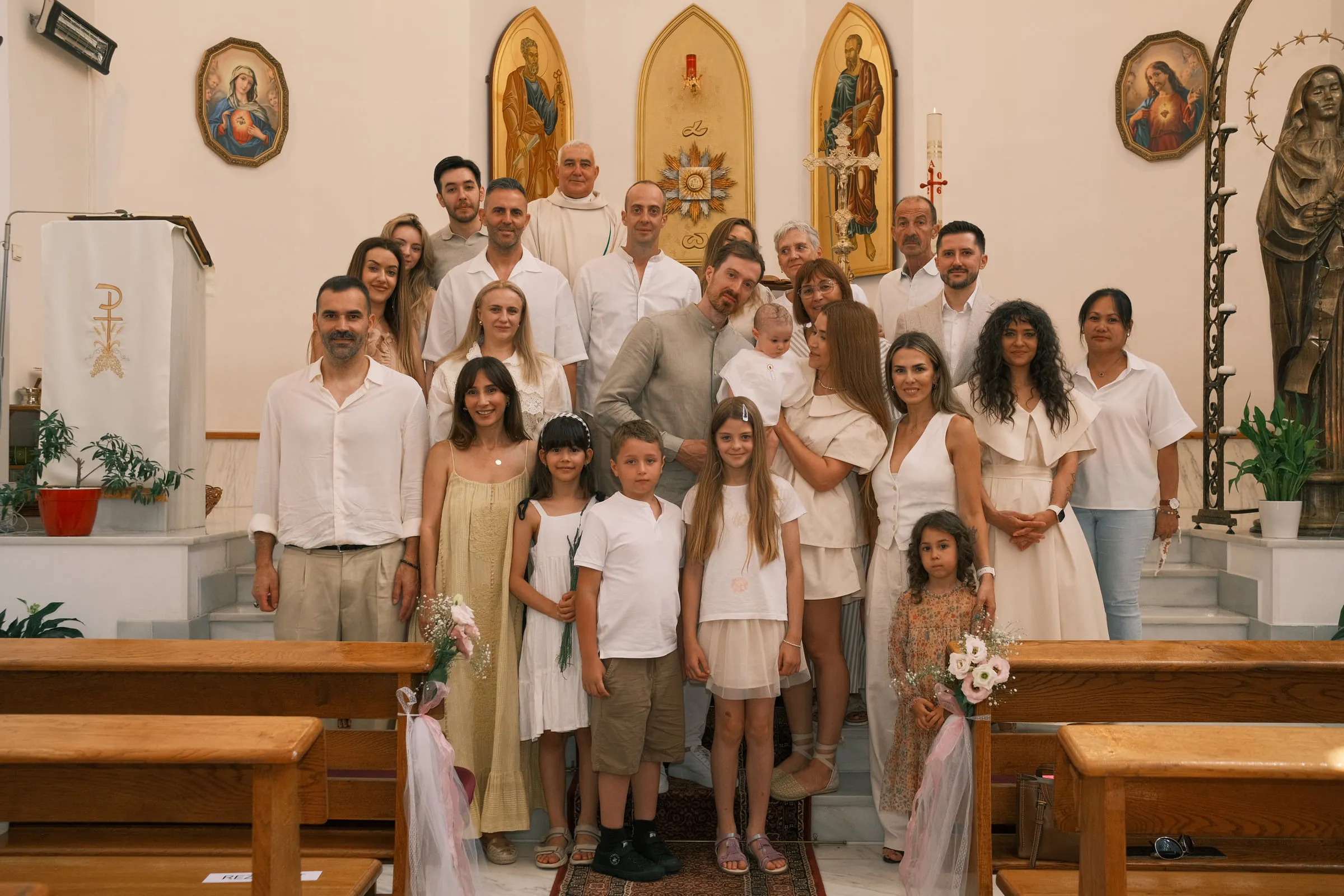 The family and guests, dressed in white and cream, gather for a group portrait before the church iconostasis
