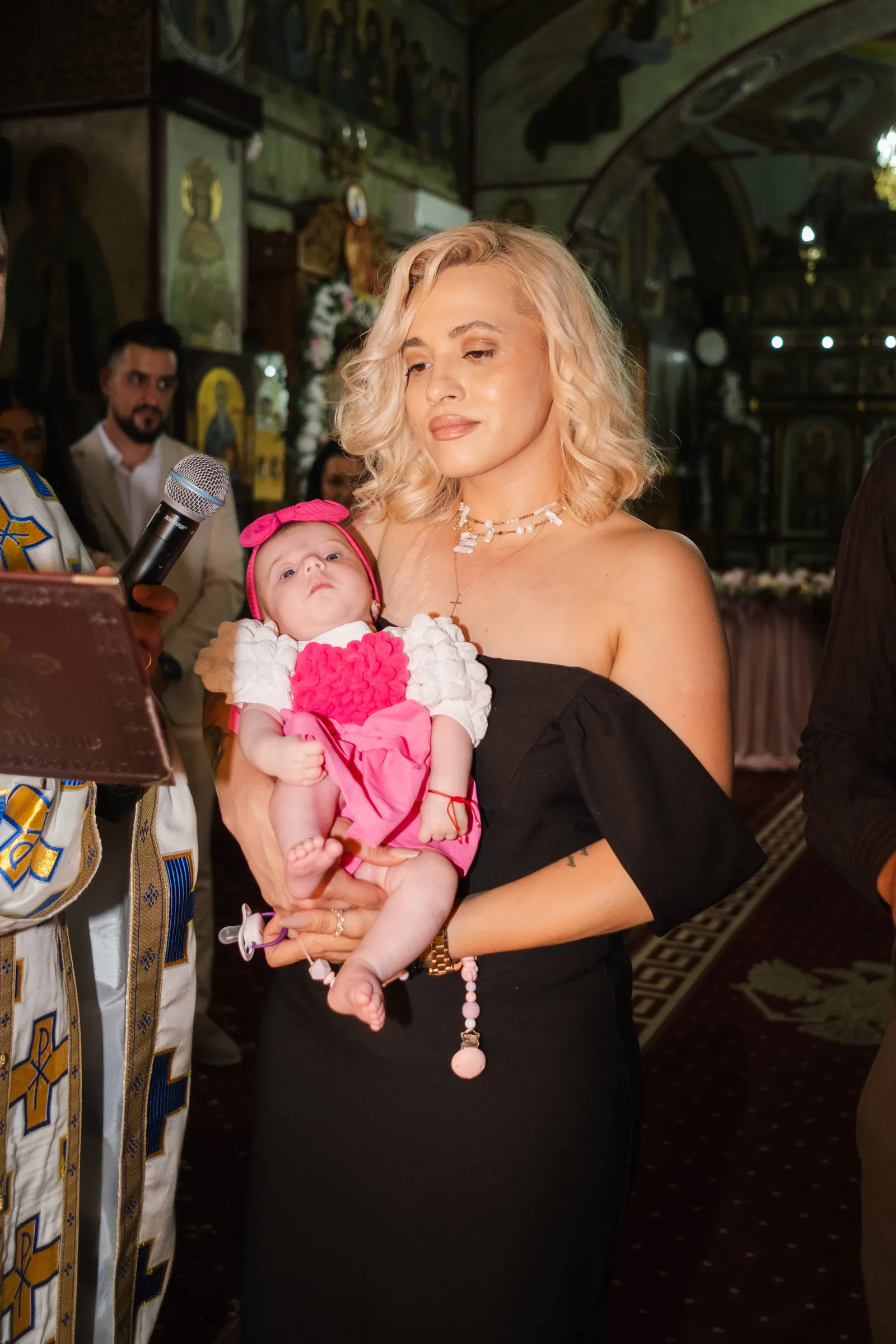 The godmother stands in the church holding the baby in pink, guests and icons behind her