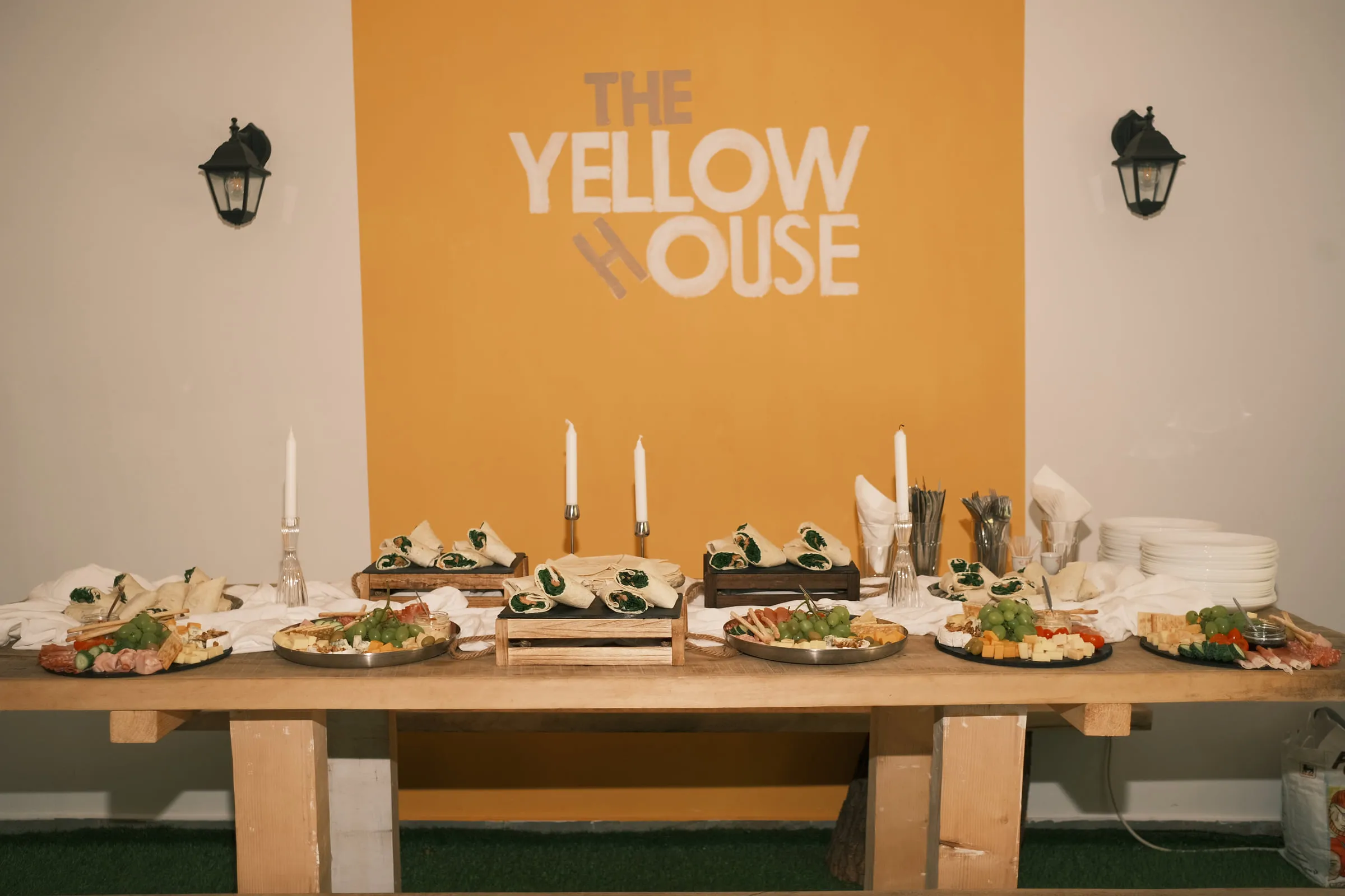 A long wooden table of food and candles set beneath The Yellow House sign