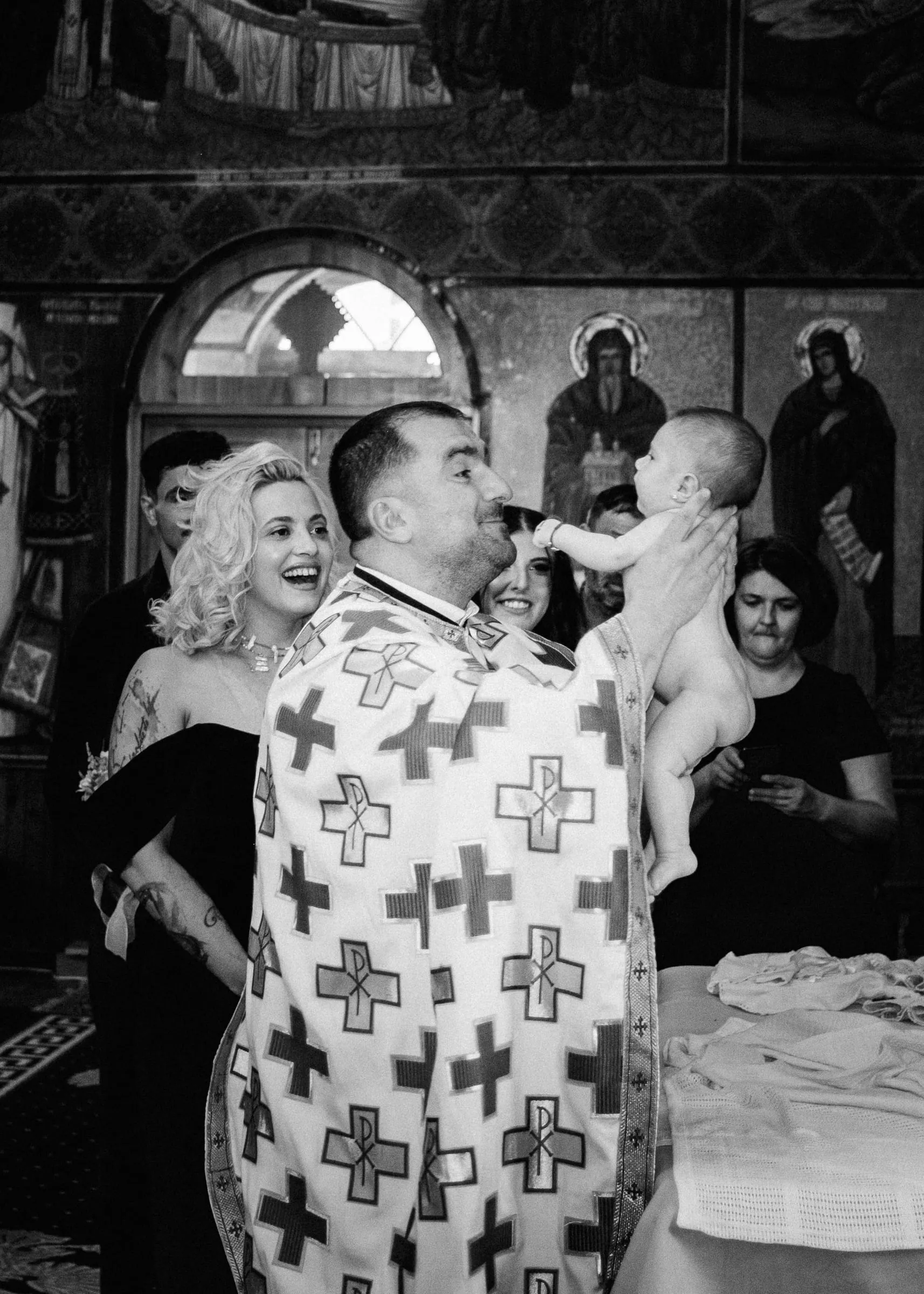 Black and white image of the priest lifting the baby high as the godmother laughs beside him