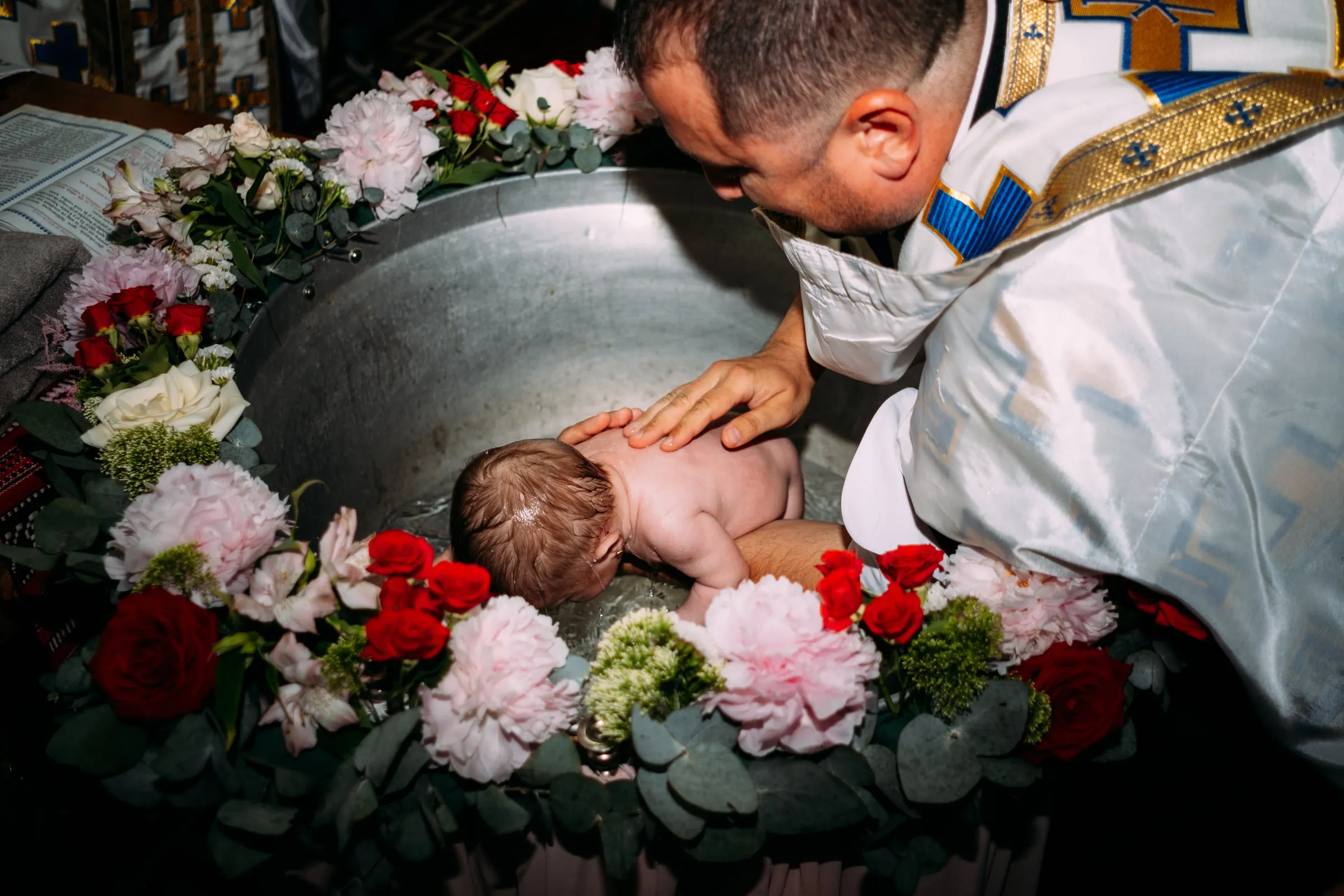 The priest gently lowers the baby into the baptismal font, its rim ringed with pink and red flowers