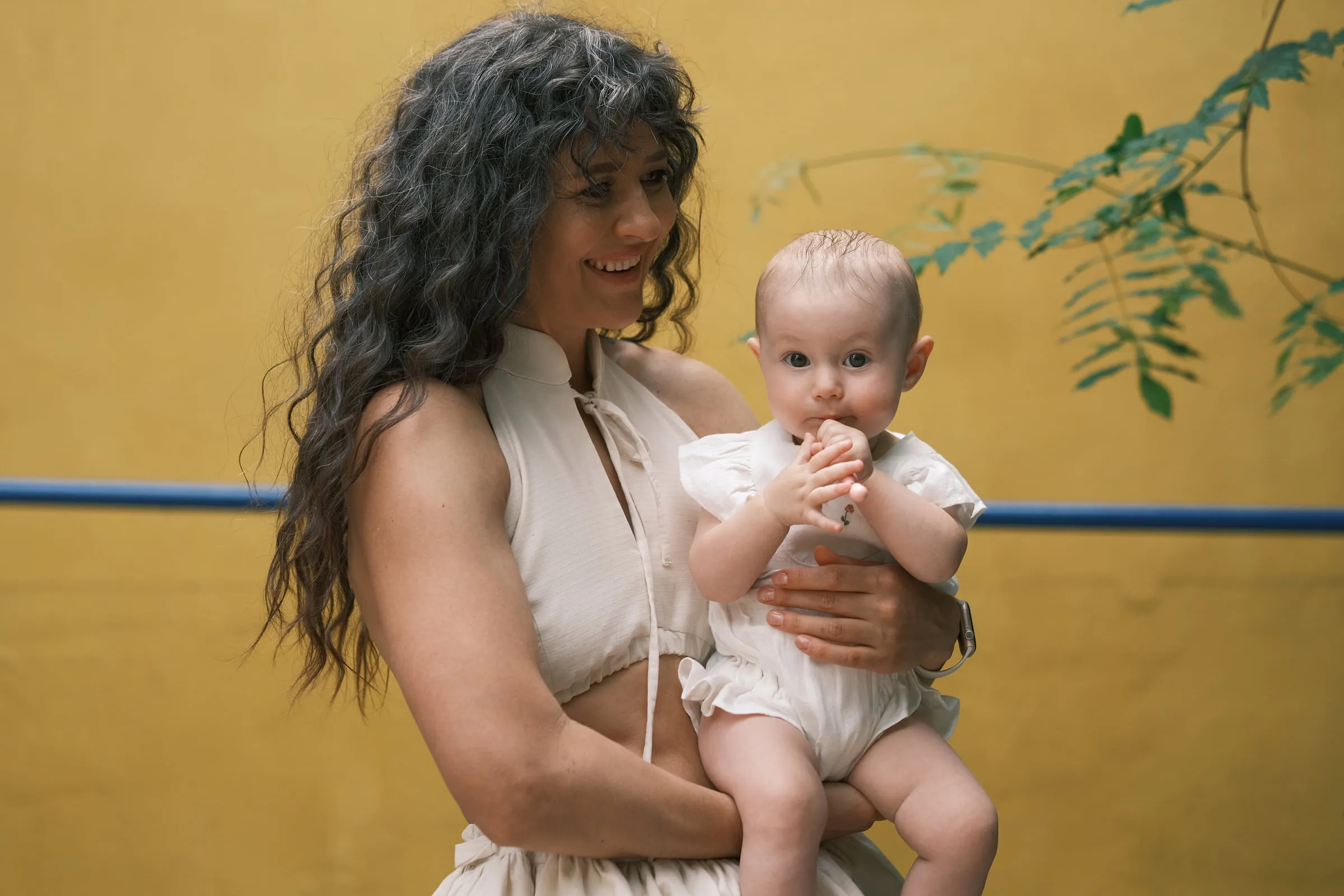 A smiling woman holds the baby against a yellow wall, leaves framing the edge of the picture