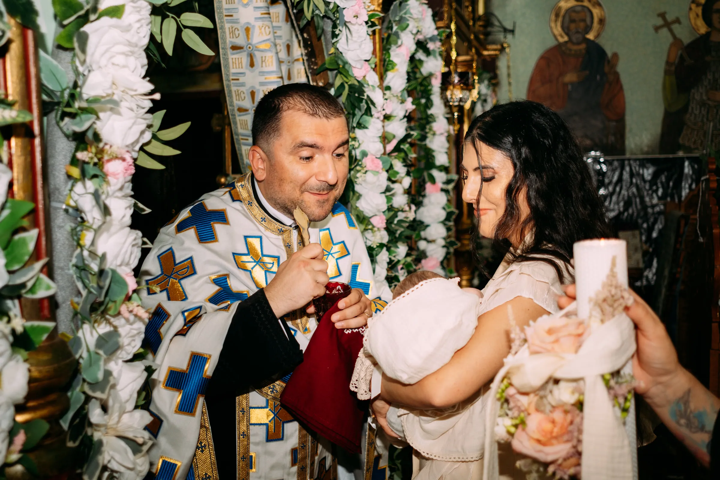 The priest smiles as he gives communion to the baby, held by her smiling mother among white flowers