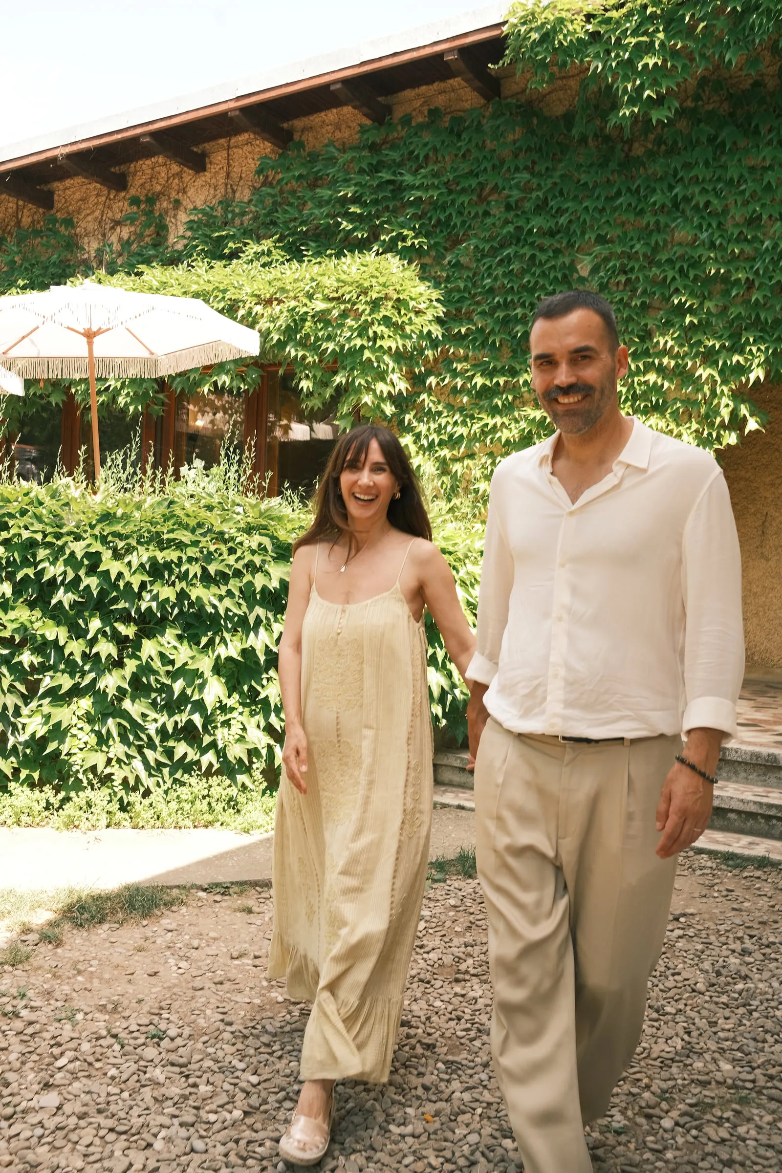 A couple walk side by side through the garden, laughing, ivy-covered building behind them